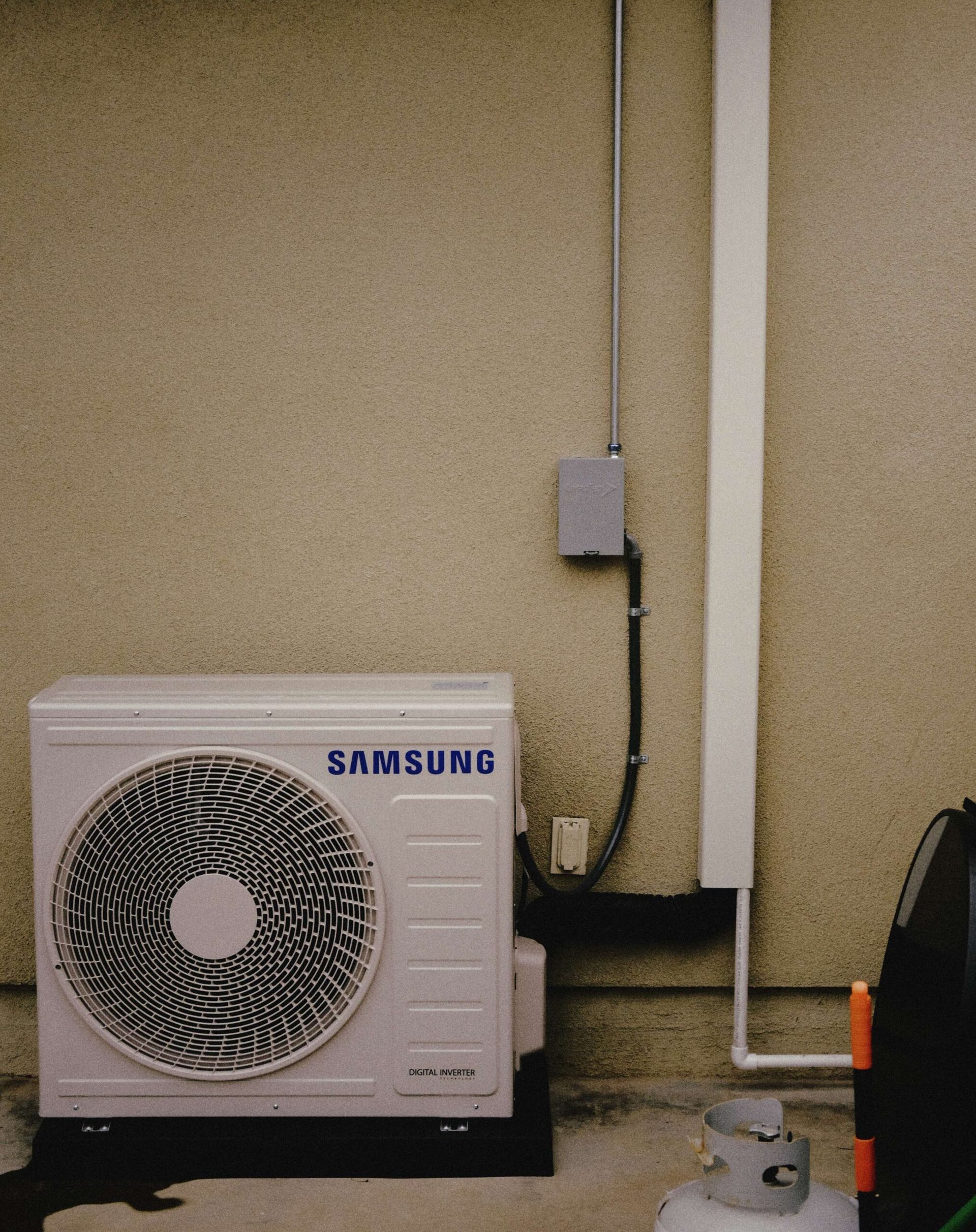 Samsung air conditioning unit against a beige wall, with electrical conduit and a propane tank nearby.