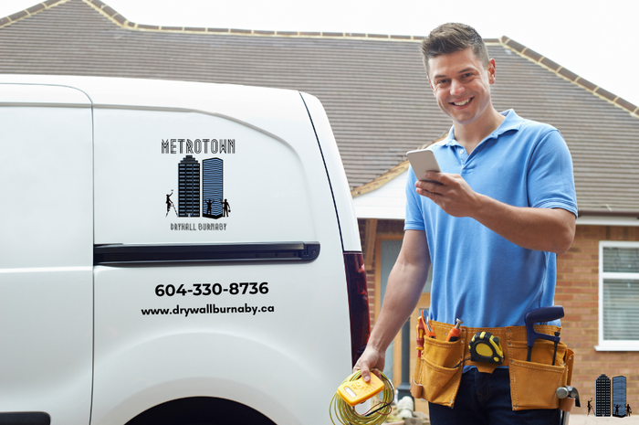 A smiling man, who appears to be a drywall contractor in Burnaby, BC, is standing next to a white van and looking at his smartphone, seemingly talking with a customer. He is wearing a blue polo shirt and a tool belt, and he holds a yellow measuring tool in his hand, suggesting he is ready for a drywall repair or drywall installation job.
