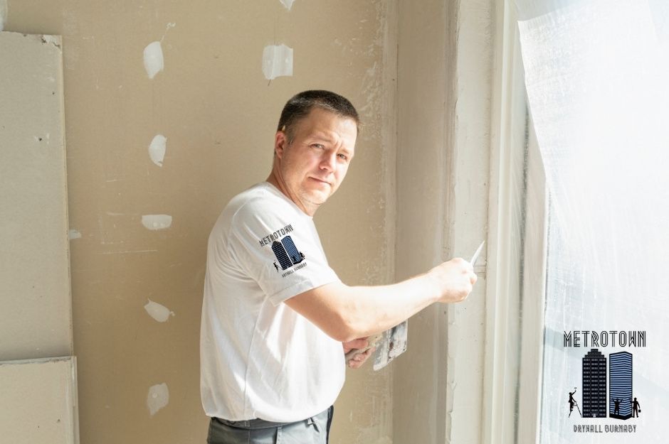 A drywall repair technician applies joint compound to seams using a putty knife during a commercial project. He wears a white shirt while working on the unfinished drywall walls, with patch repairs visible throughout the room near W 55th Ave and Blenheim St in South Vancouver, BC.