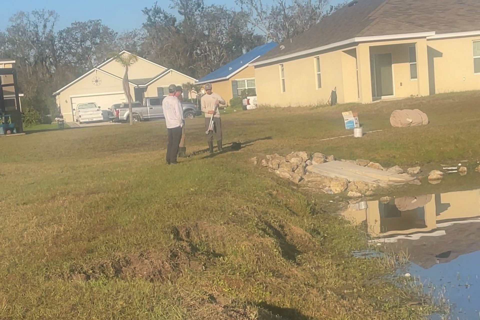 Two men stand in a grassy yard by a pond. One holds a document, houses in the background.