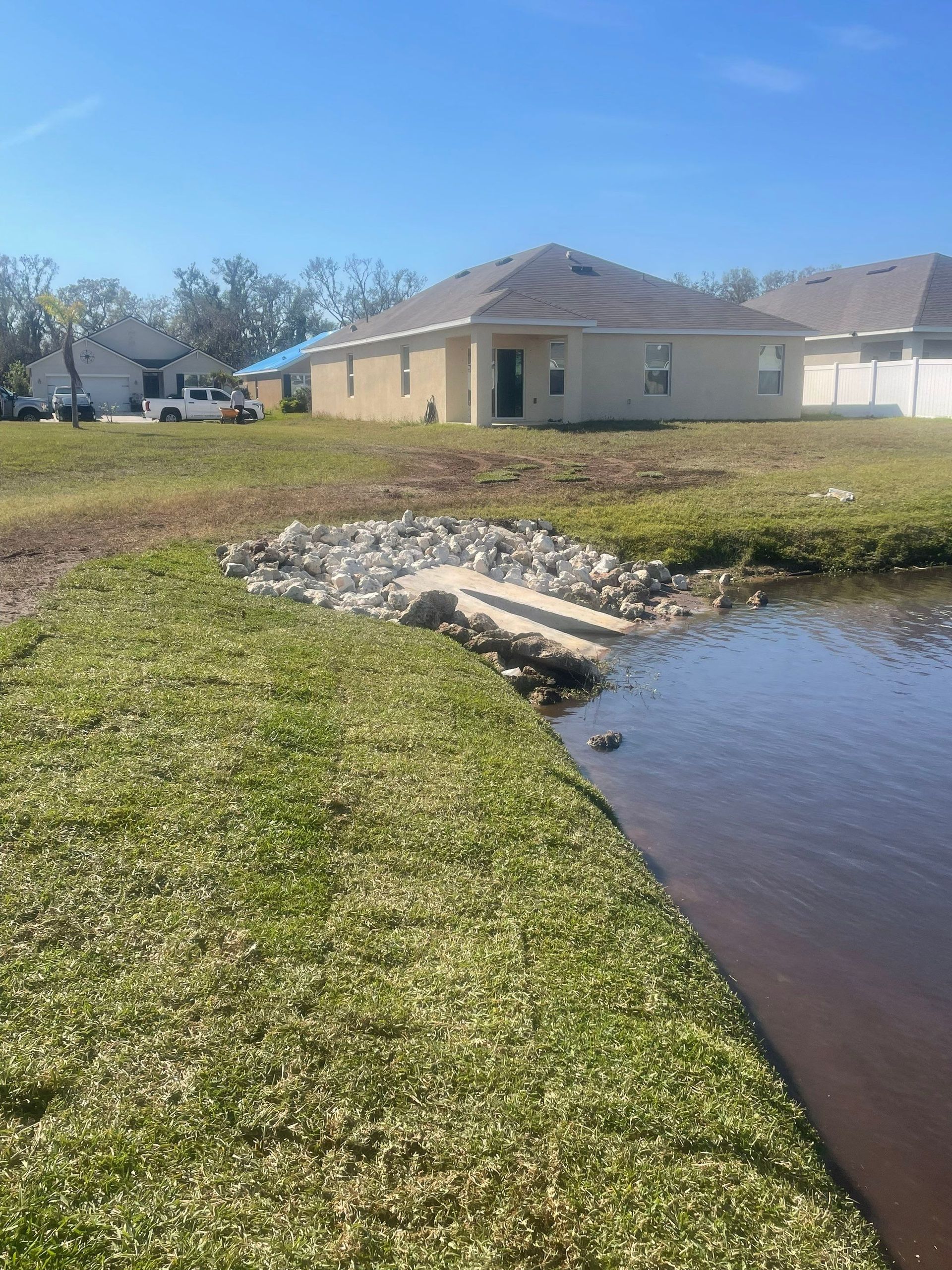Grassy bank leads to a small concrete area with rocks at the edge of a body of water, behind a light-colored house.