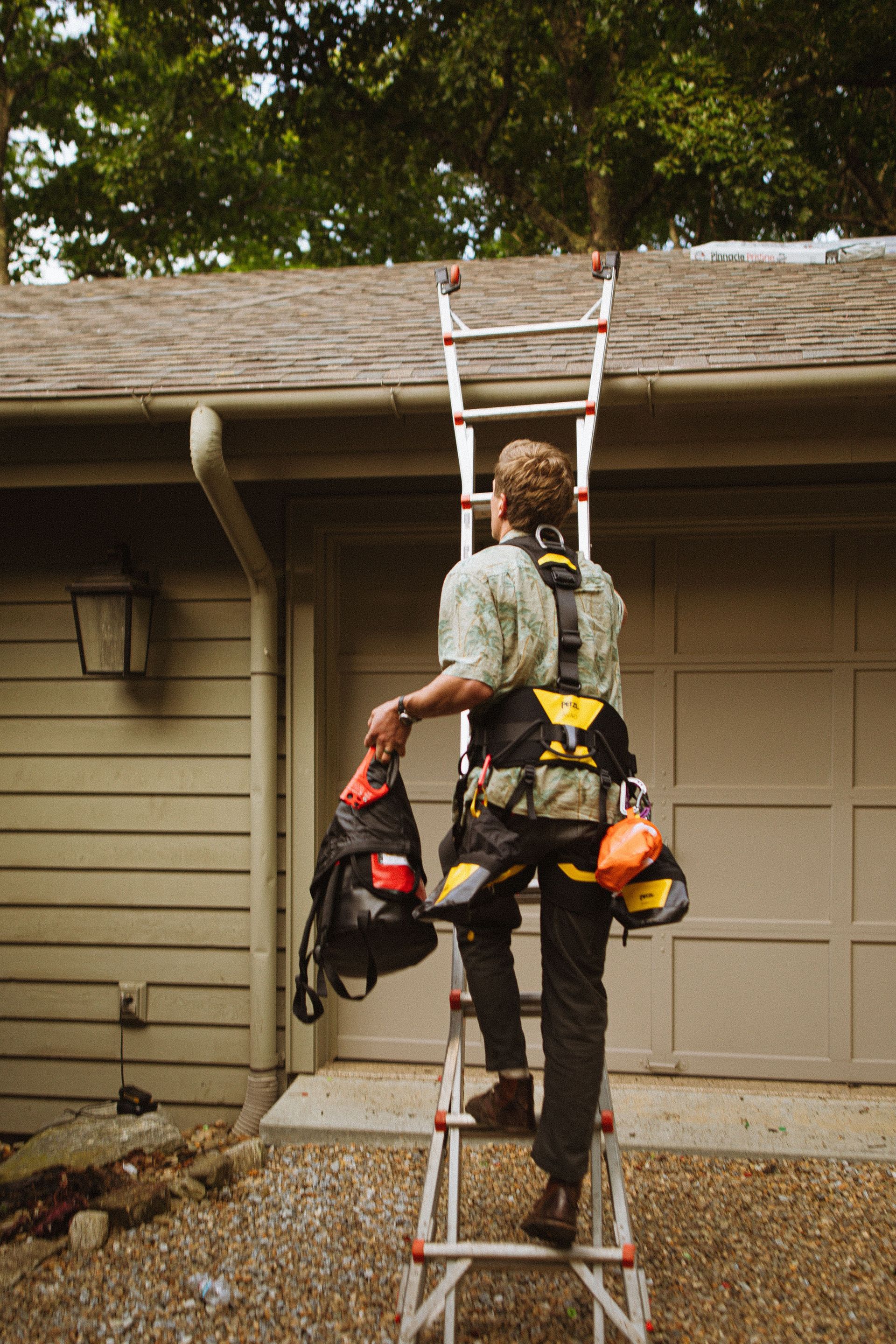 Worker on a ladder cleaning a house gutter, carrying tools on a harness.