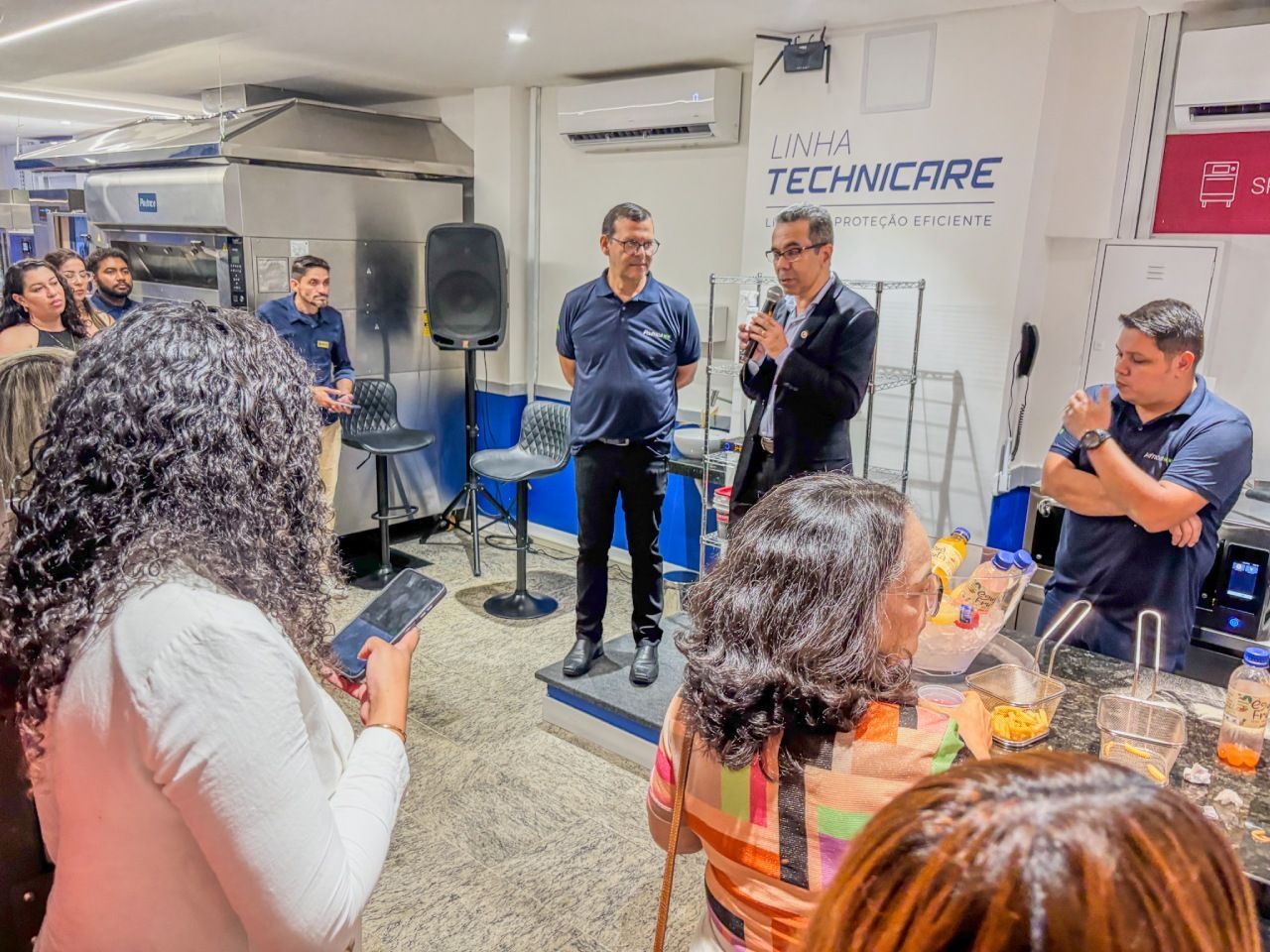 Pessoas em um evento. Um homem fala ao microfone. Outras pessoas ouvem.