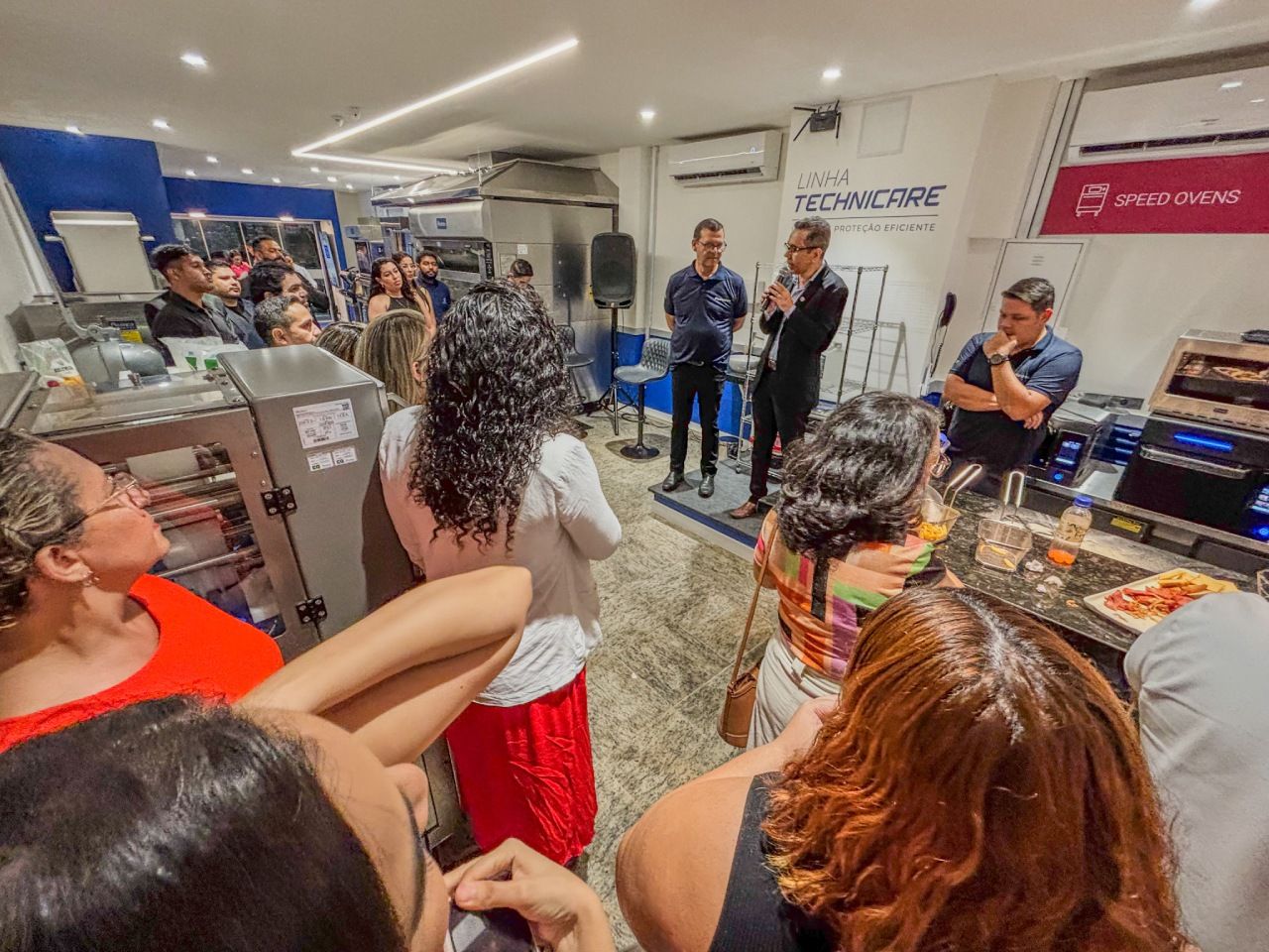 Pessoas em um evento, dois homens conversando. Foto de interior: decoração azul e branca, comida e equipamentos visíveis.