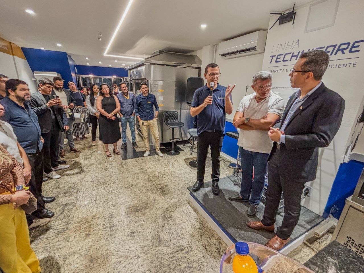 Grupo de pessoas em uma loja de eletrônicos, dois homens conversando em uma plataforma elevada, outros ouvindo, esquema de cores azul claro e branco.