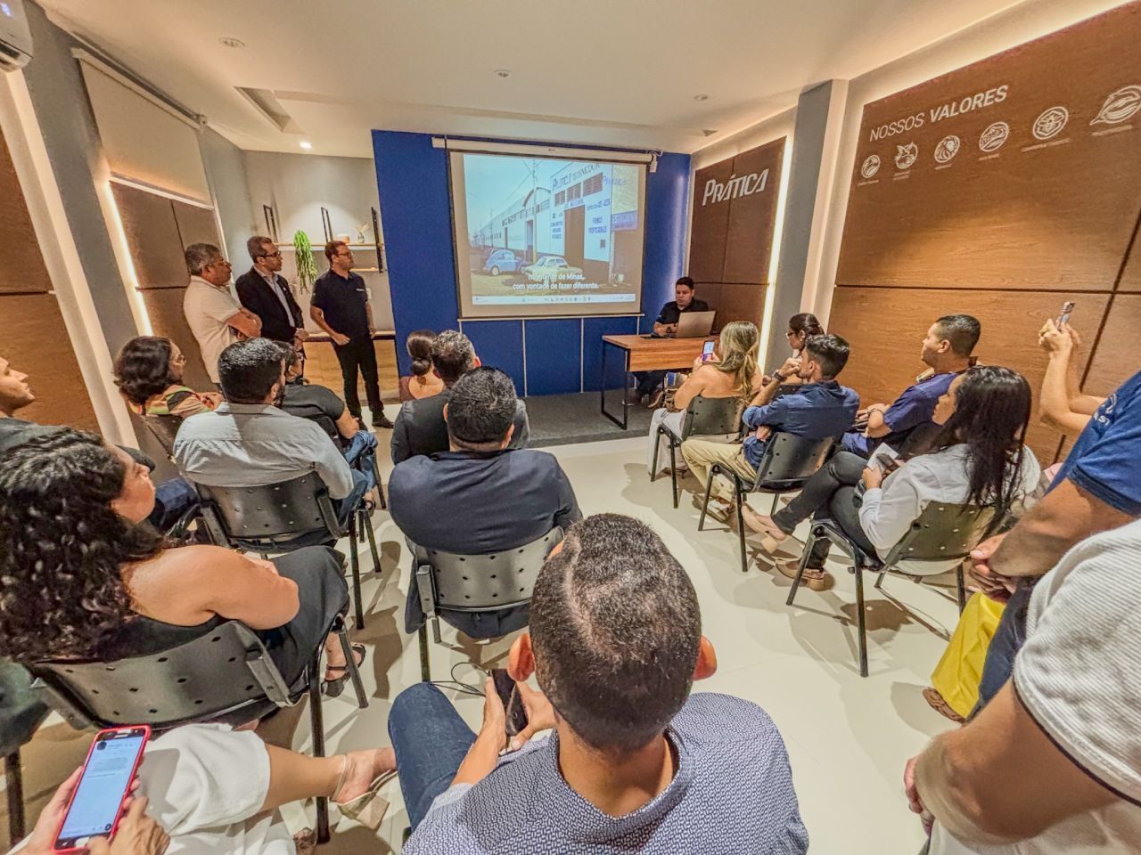 O público assiste à apresentação em uma sala com uma tela. Dois homens estão em pé ao lado dela; um sentado à mesa.