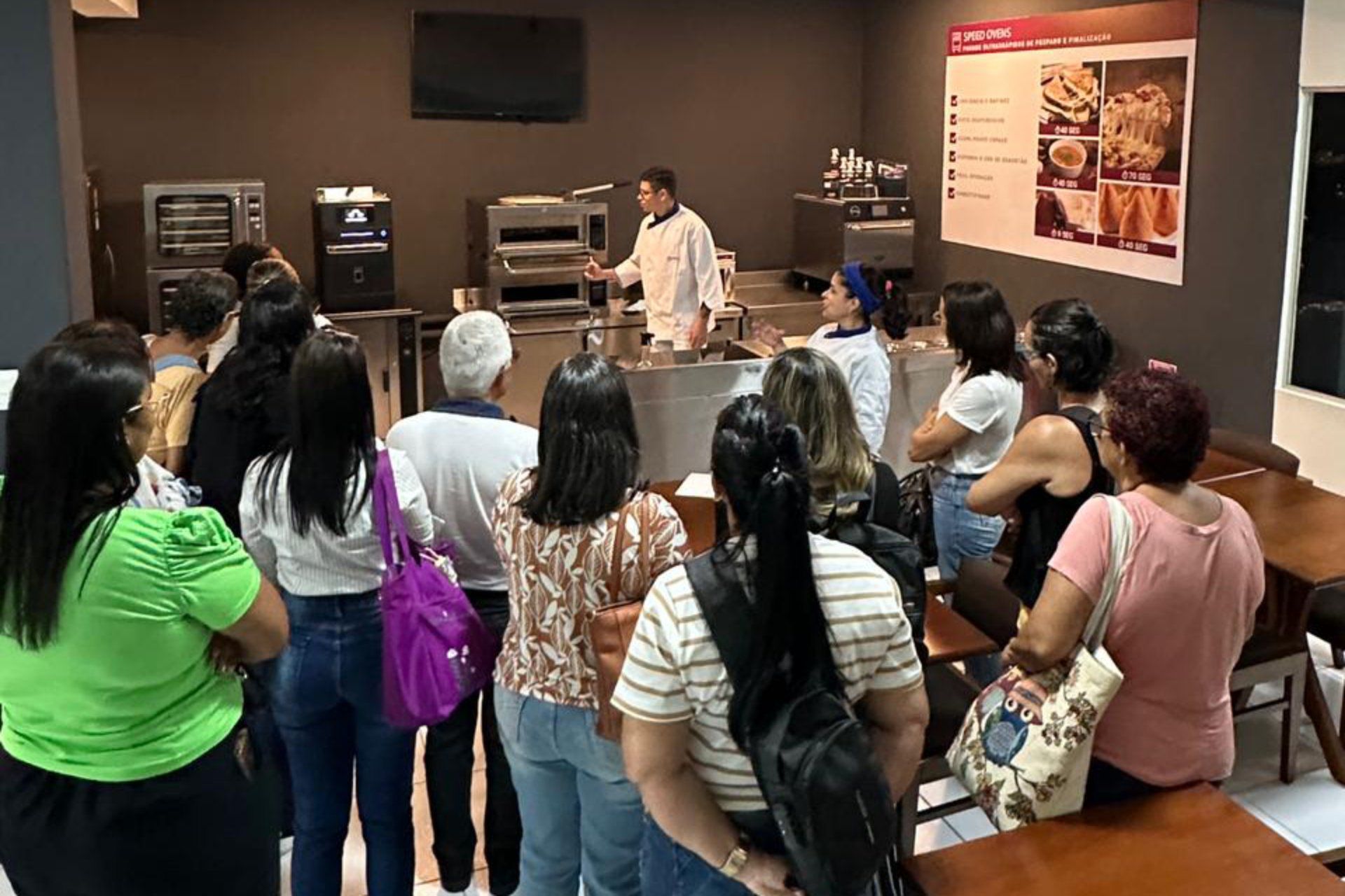 Um grupo de pessoas está em uma sala observando um homem cozinhar.