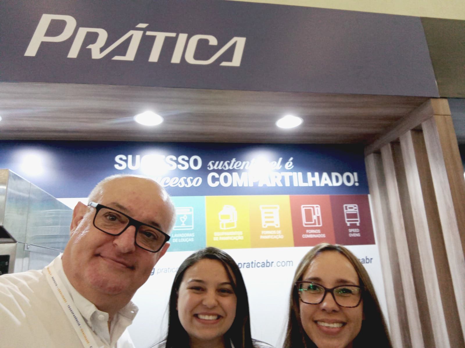 Three people smiling in front of a Prática sign, which also features the phrase