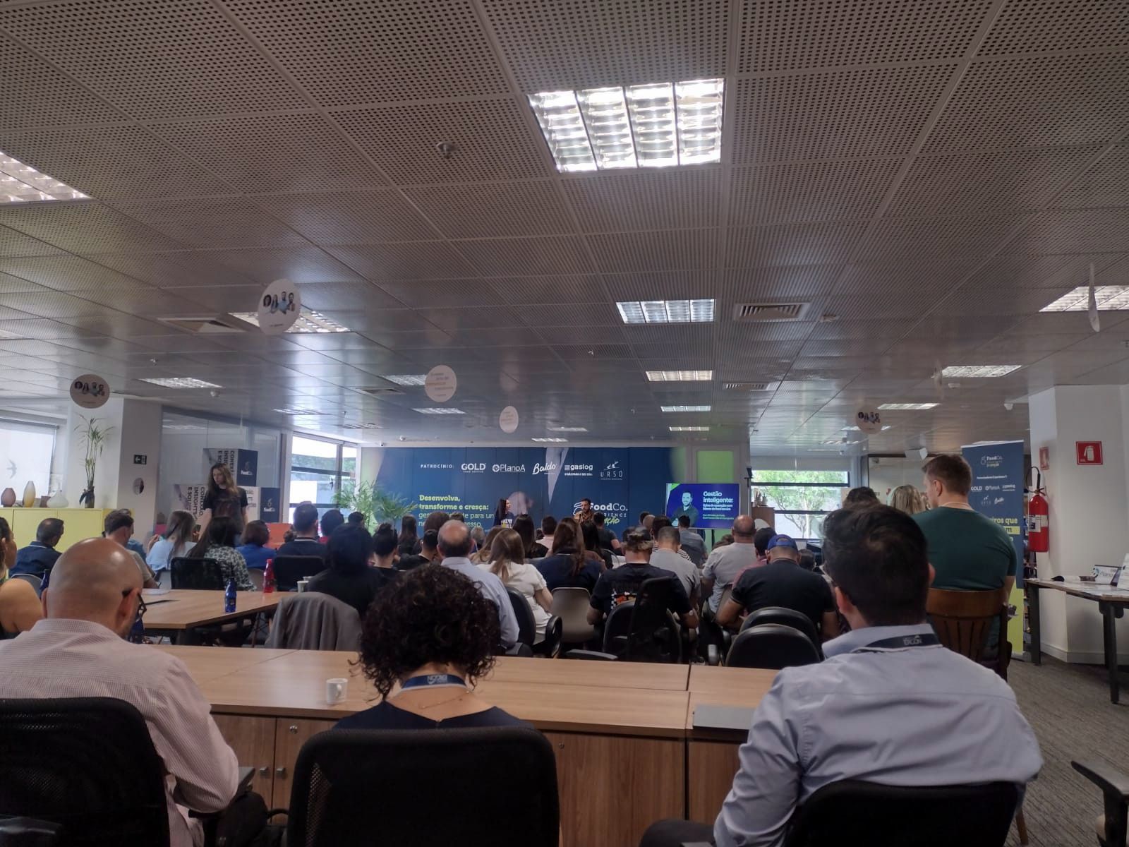 Um grande grupo de pessoas está sentado em uma sala com mesas e cadeiras.