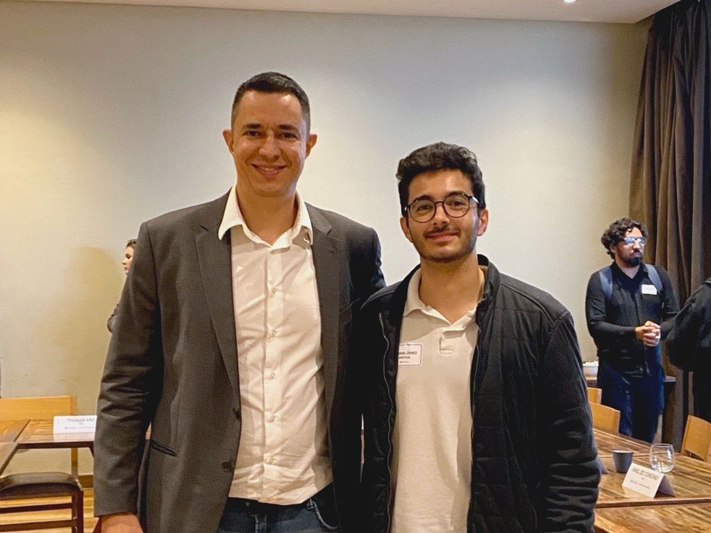 Dois homens sorrindo em uma sala; um de blazer, o outro de jaqueta preta. Outras pessoas ao fundo.