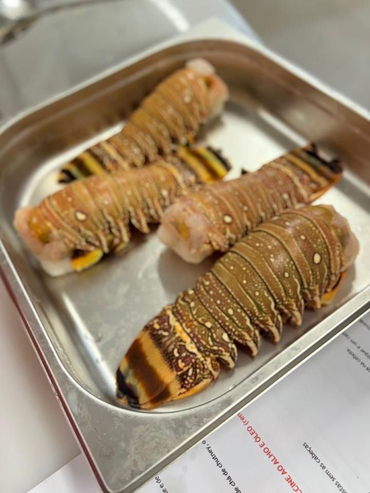 Quatro caudas de lagosta cruas em uma bandeja de metal, mostrando caudas listradas e carne laranja clara.