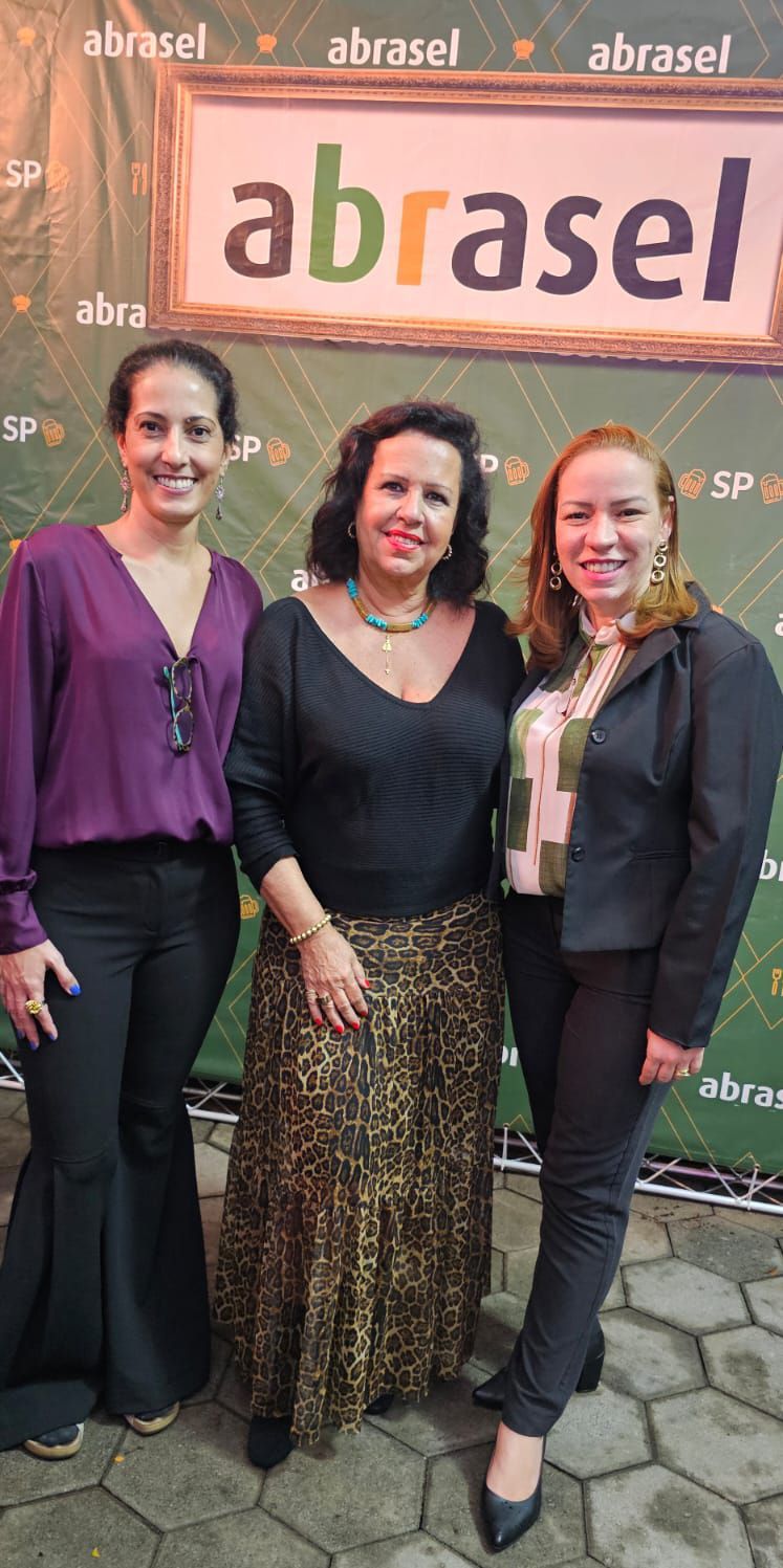 Três mulheres estão posando para uma foto em frente a uma placa que diz abrasel.