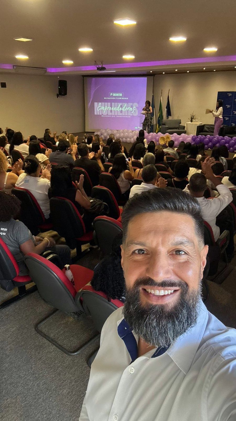 Homem tira selfie em um auditório com uma apresentação em uma tela e uma grande plateia.