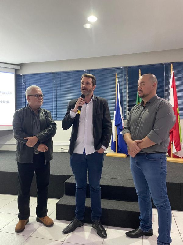Três homens estão em pé na frente de bandeiras e um está falando em um microfone