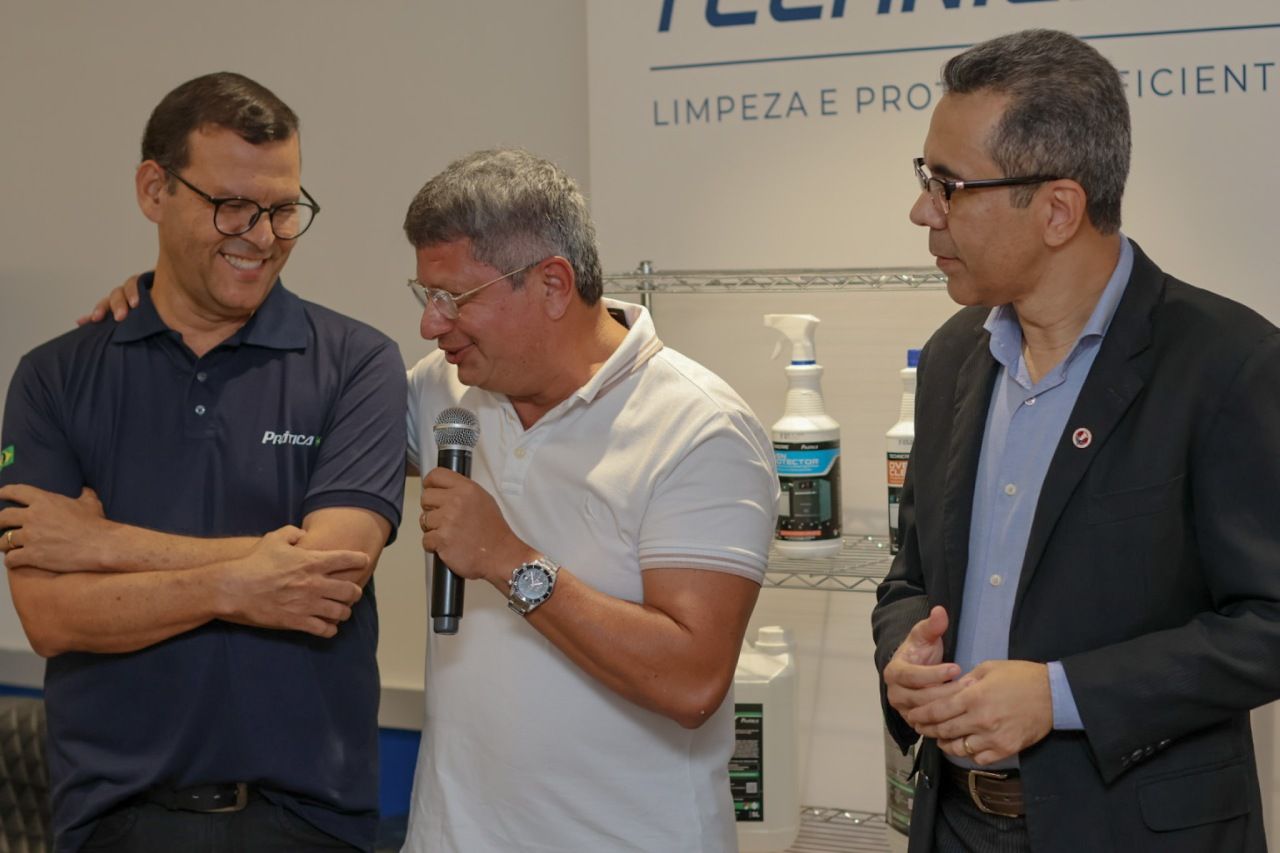Três homens em um evento. Um fala ao microfone, outro tem o braço em seu ombro e o terceiro observa.