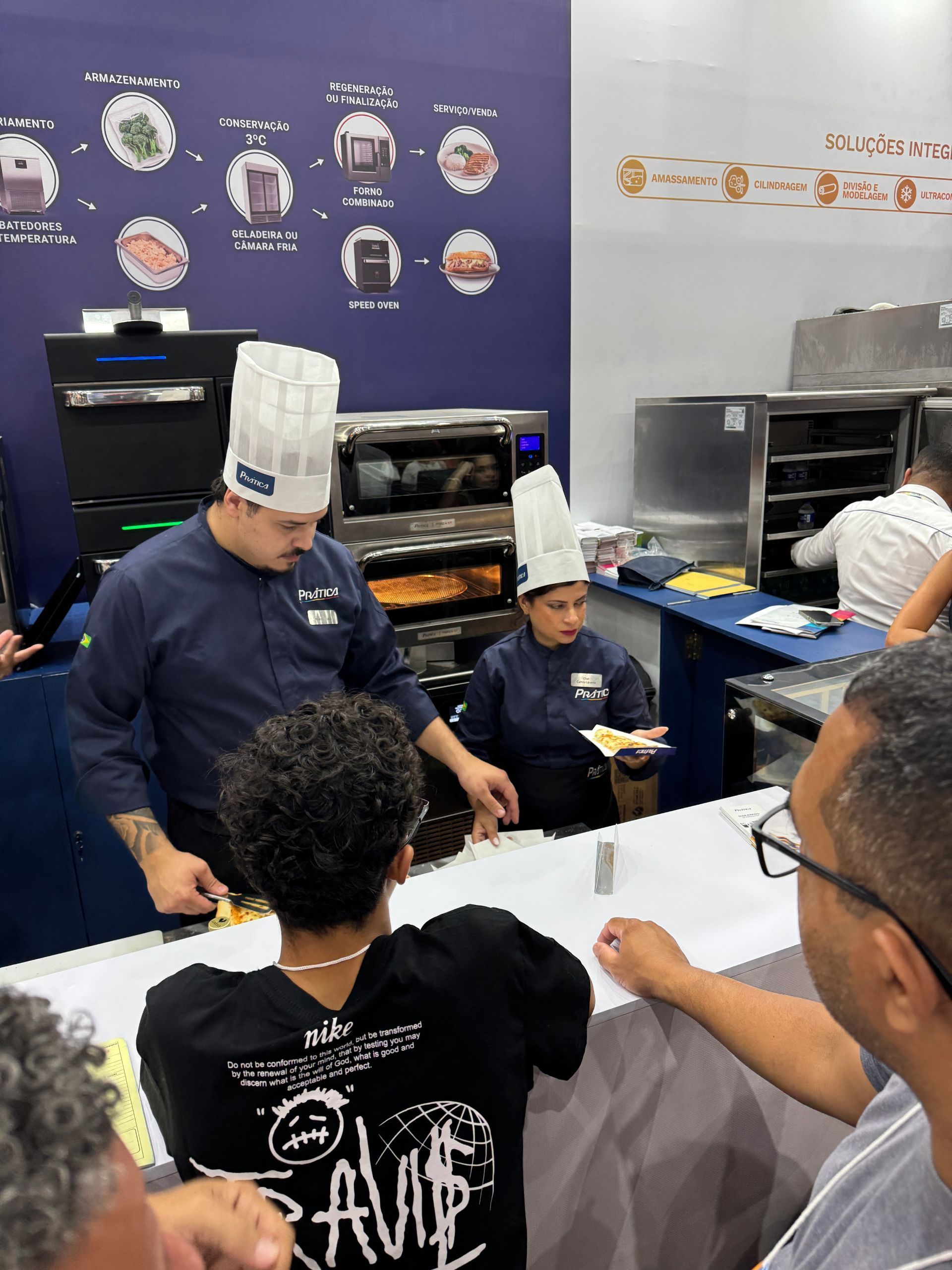 Chefs uniformizados preparando comida atrás de um balcão, com uma multidão de pessoas assistindo.
