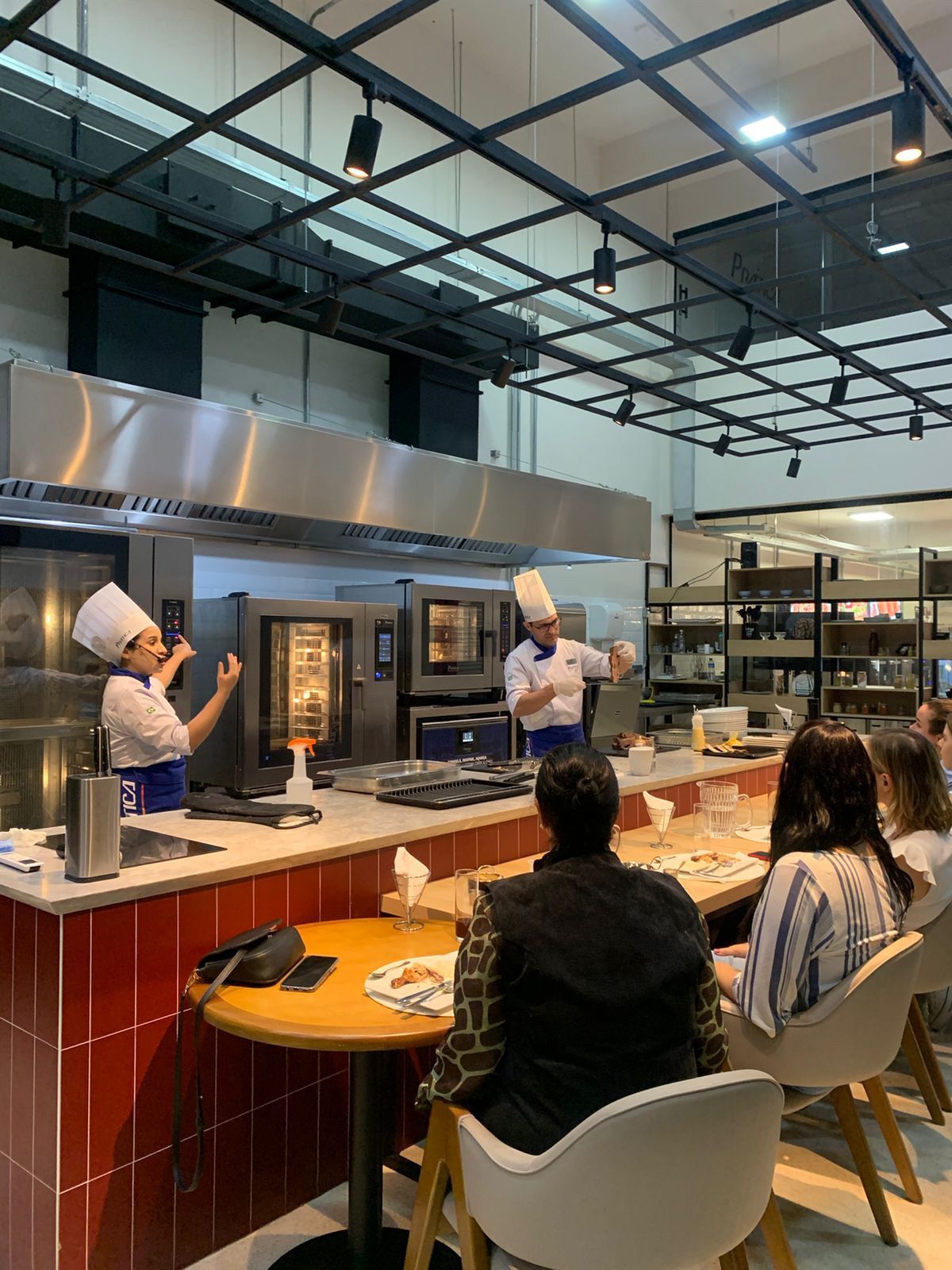 Um grupo de pessoas está sentado em mesas em uma cozinha observando dois chefs cozinhando.
