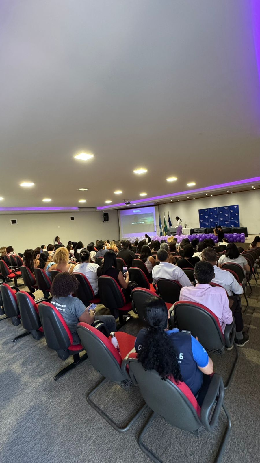 Público em uma sala de conferências; um palestrante está na frente, a apresentação é exibida em uma tela.