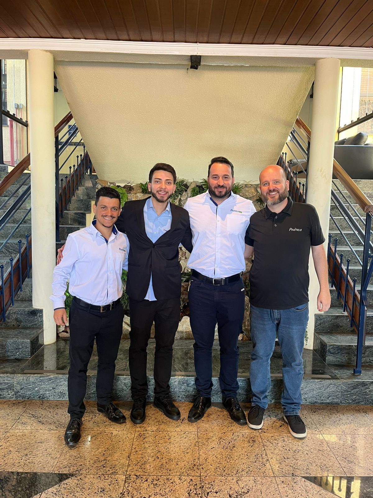 Quatro homens posam para uma foto em um ambiente fechado. Um veste uma camisa polo preta e calça jeans; os outros estão em trajes formais.