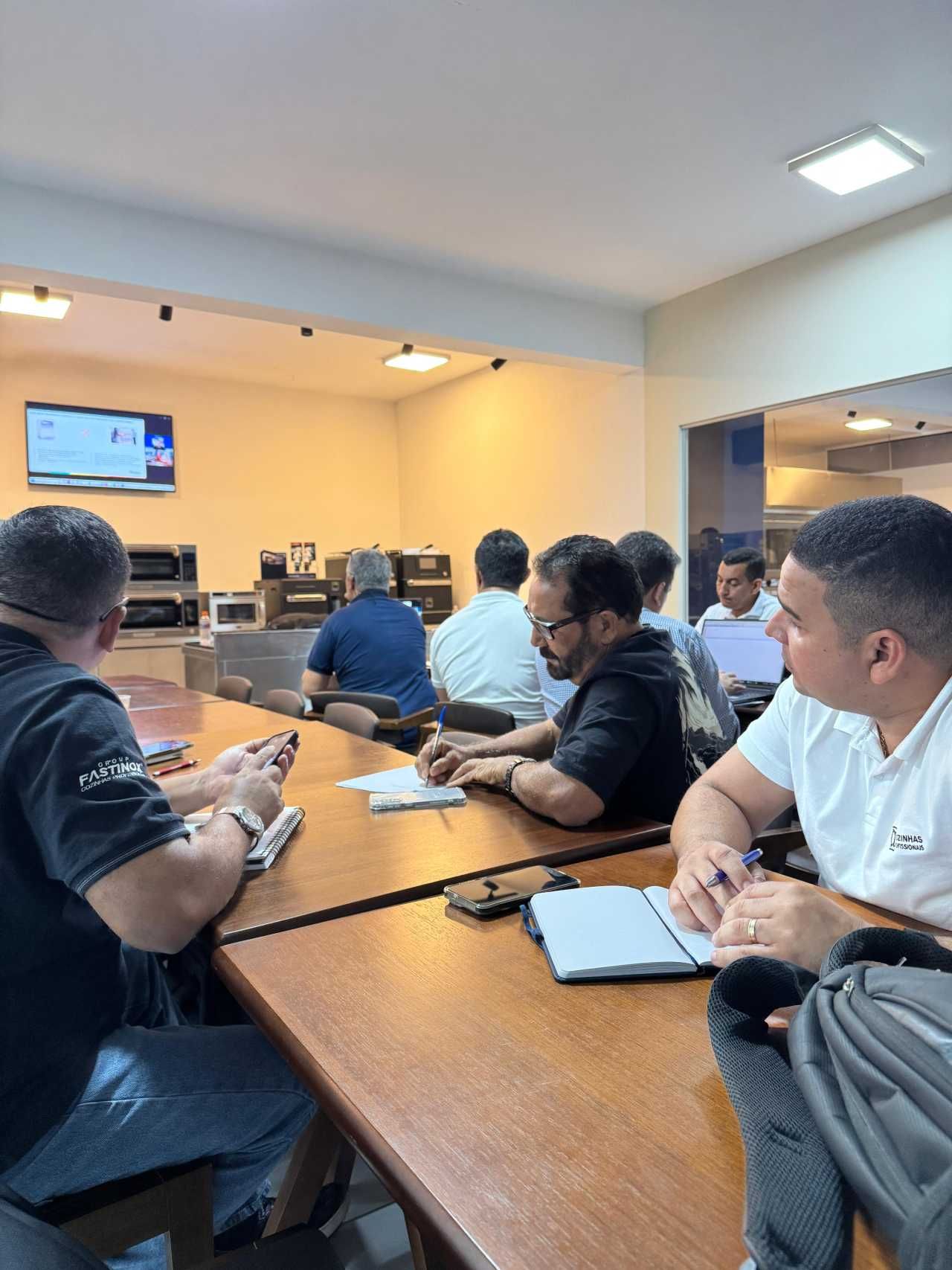 Em uma sala bem iluminada, pessoas sentam-se em torno de uma longa mesa de madeira, olhando para cadernos. Uma TV exibe um noticiário.