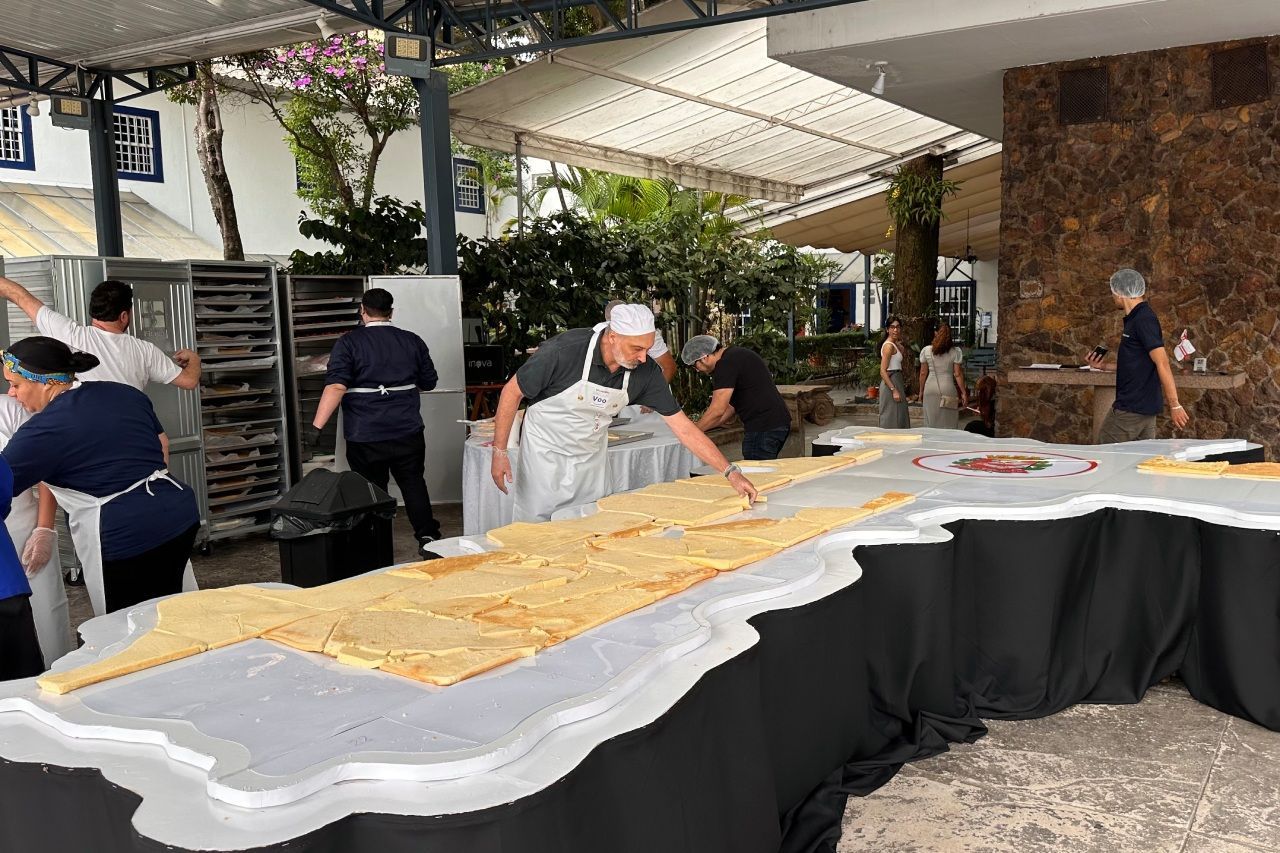 Pessoas preparando um alimento grande e cortado em pedaços sobre uma mesa. Ao ar livre, aventais brancos e produtos assados.