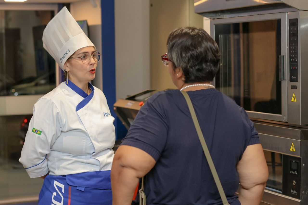 Um chefe de cozinha uniformizado conversa com uma pessoa perto dos fornos em uma cozinha industrial.