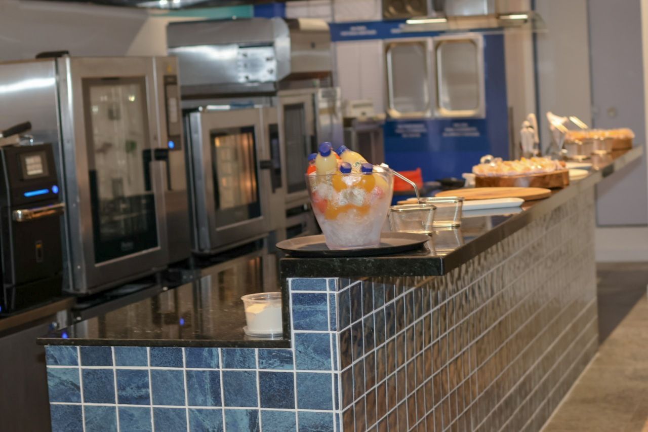 Uma cozinha comercial com fornos e uma bancada com sobremesas e uma salada de frutas.