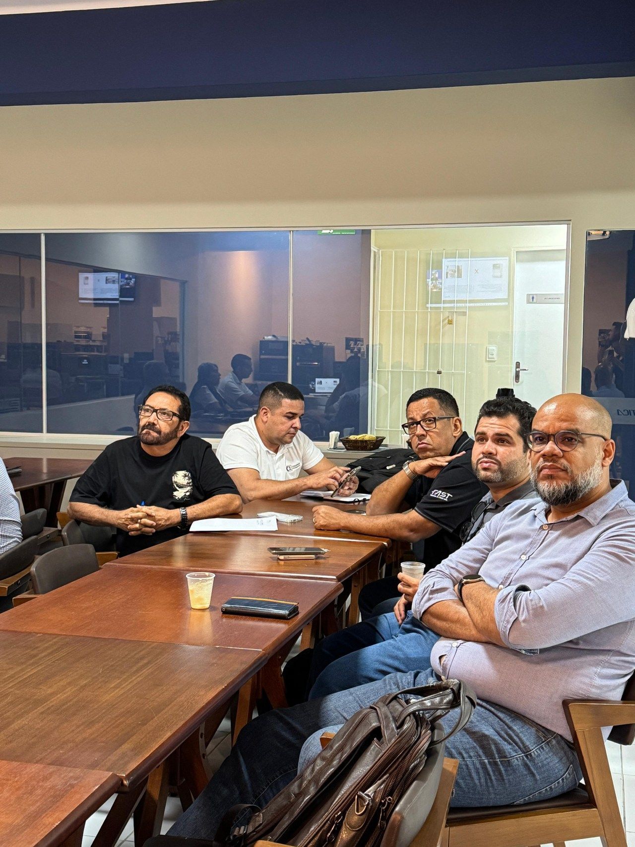 Homens sentados a uma mesa de madeira em uma sala com janela, aparentemente em reunião.