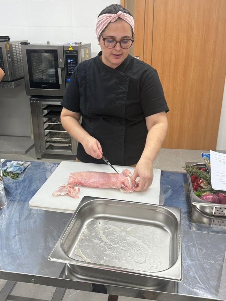 Chef de uniforme preto, cortando carne em uma tábua branca, área de preparação de aço inoxidável.