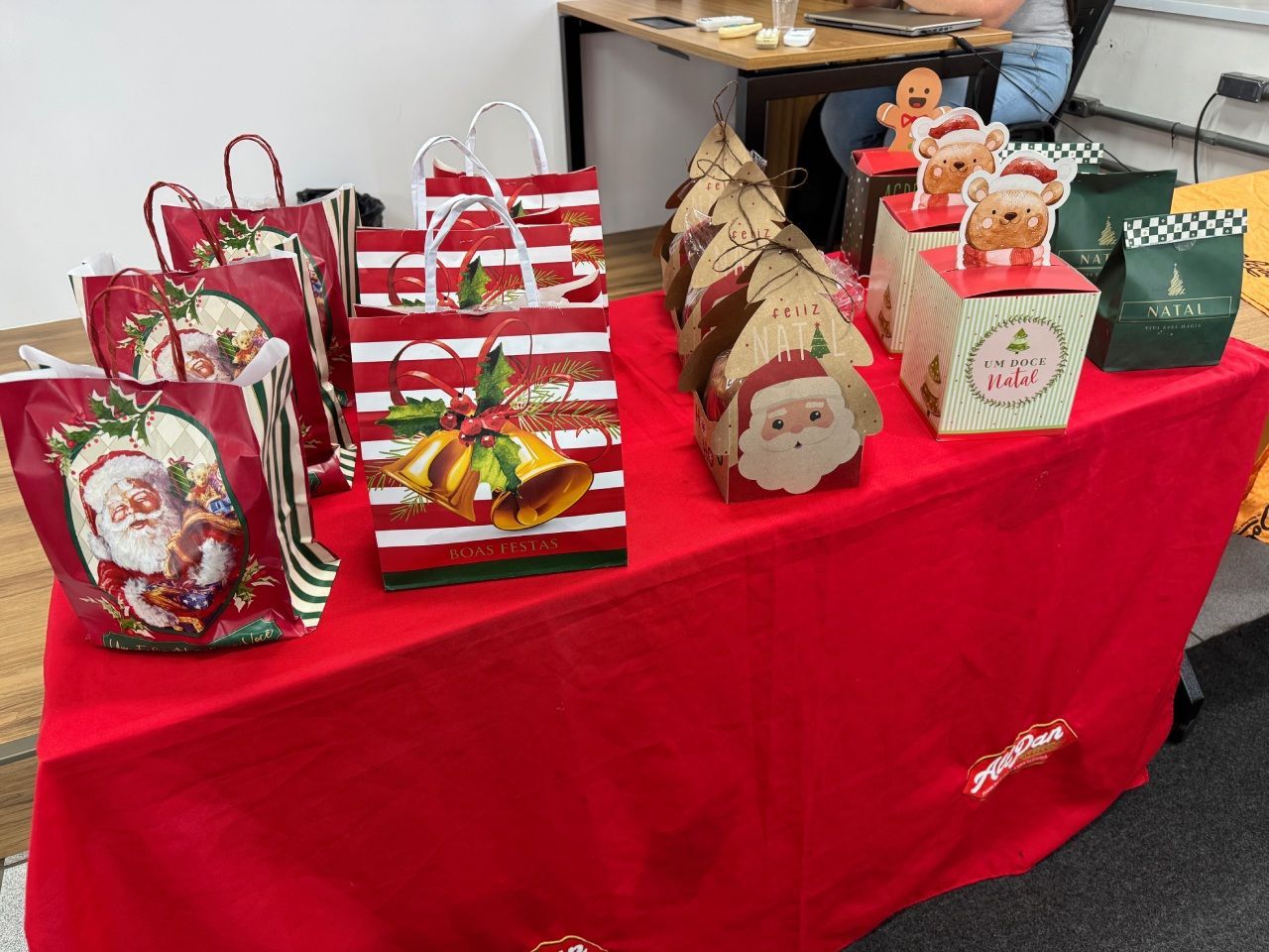 Sacolas e caixas de presentes de Natal sobre uma mesa vermelha. Papai Noel, sinos e árvores decoram as embalagens.
