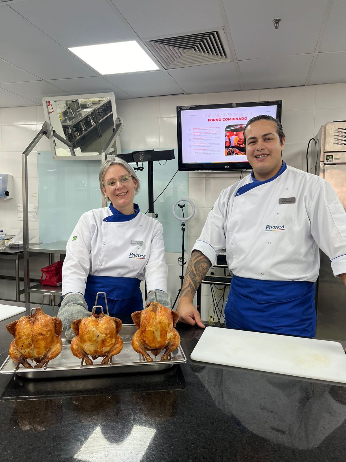 Um homem e uma mulher estão de pé ao lado de uma bandeja de frangos em uma cozinha.