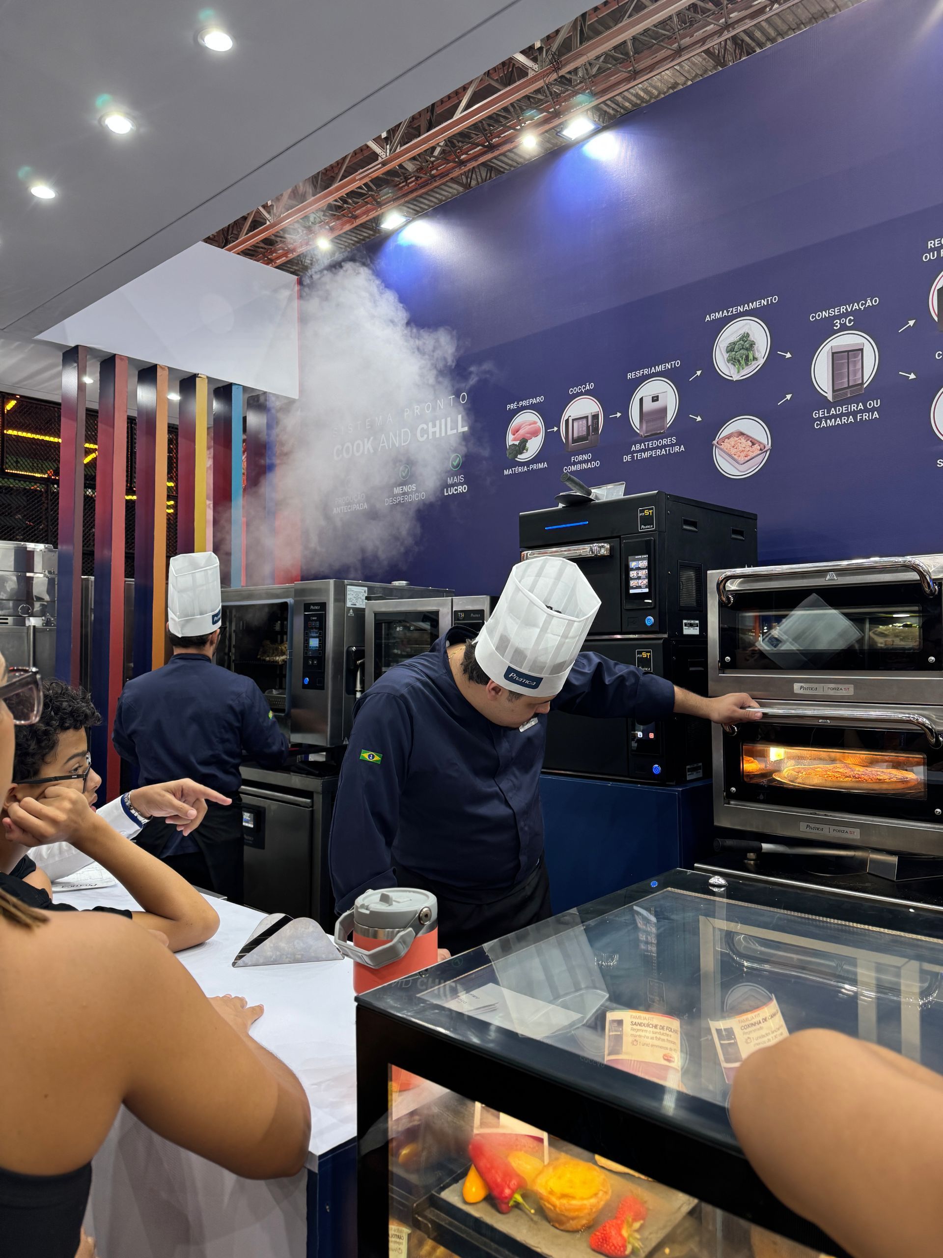 Chefs de chapéu branco e uniforme azul operam fornos com vapor visível em uma barraca de feira gastronômica.
