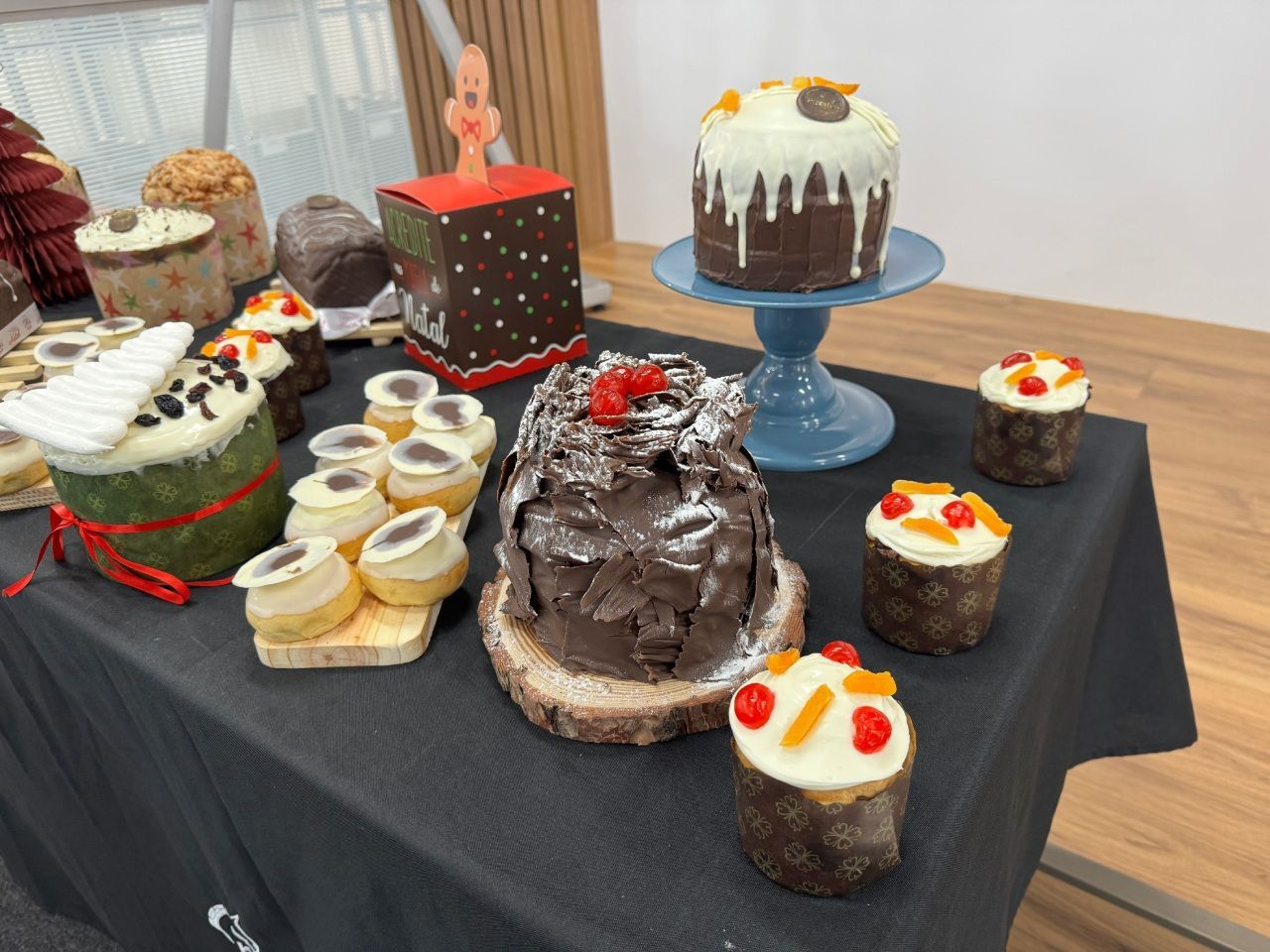 Exposição de diversos bolos decorados sobre uma mesa, incluindo cupcakes e um bolo em um suporte azul; superfície escura.