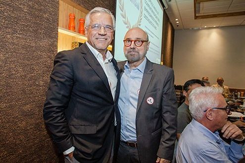 Dois homens de terno posam para uma foto em uma sala com uma tela de projeção. Um deles está com a mão no ombro do outro.
