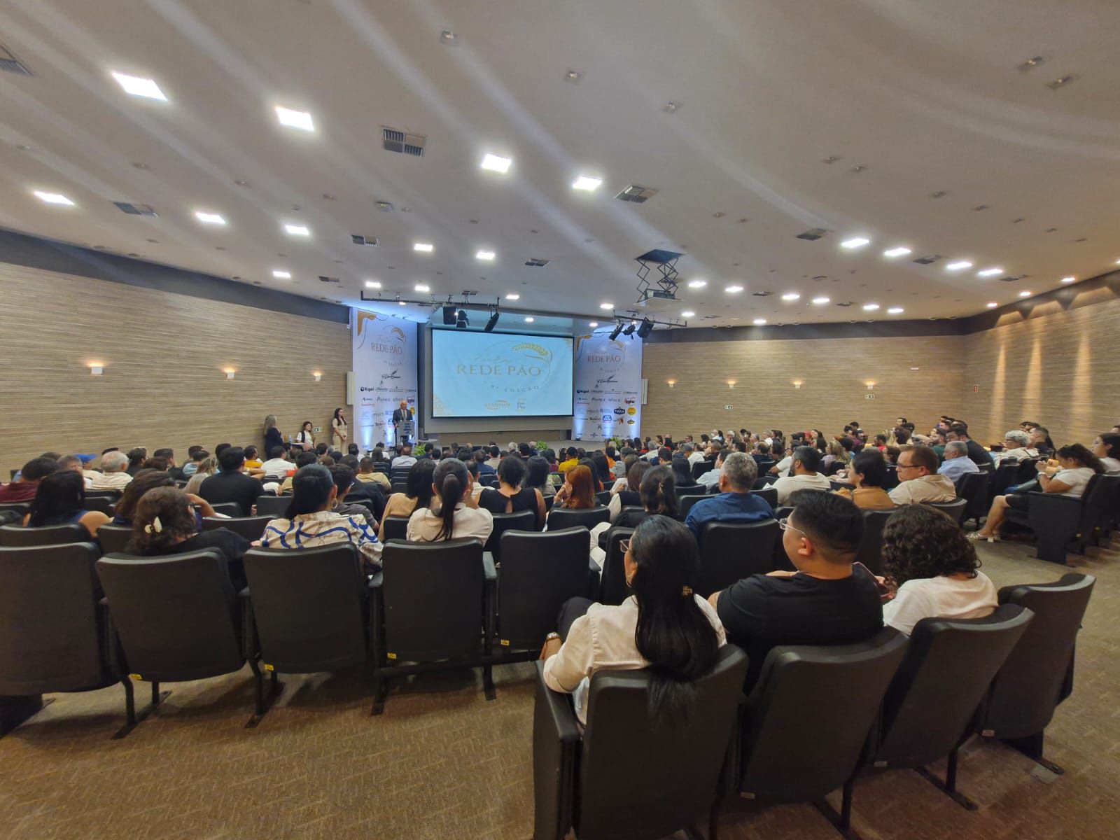 Um grande auditório cheio de pessoas sentadas em cadeiras assistindo a uma apresentação.