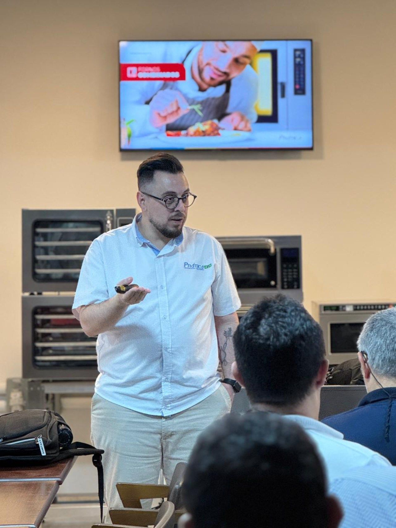 Homem fazendo uma apresentação em uma cozinha, apontando para a plateia. Tela de TV e fornos industriais ao fundo.
