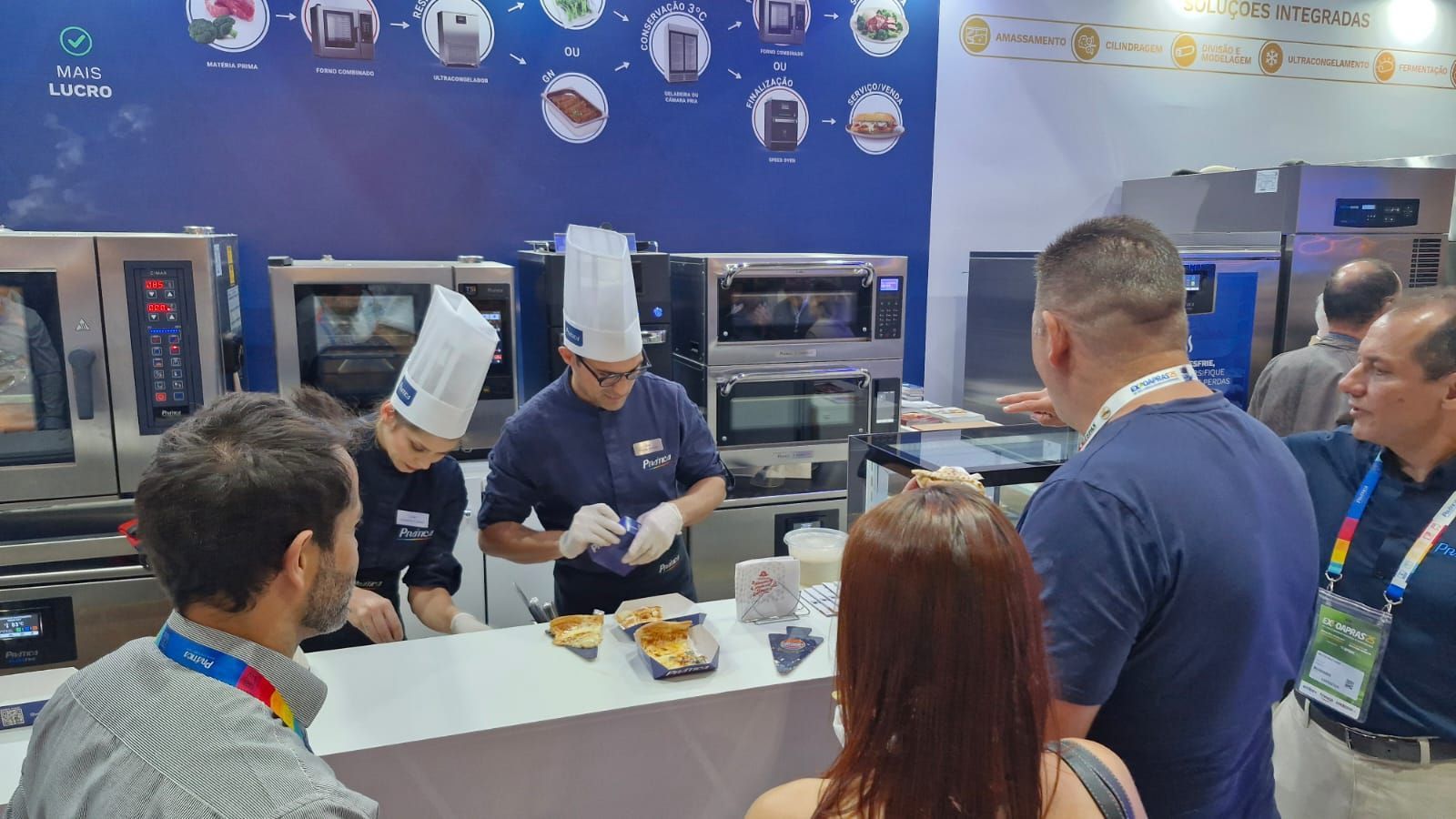 Um grupo de pessoas está em pé ao redor de uma mesa na cozinha.