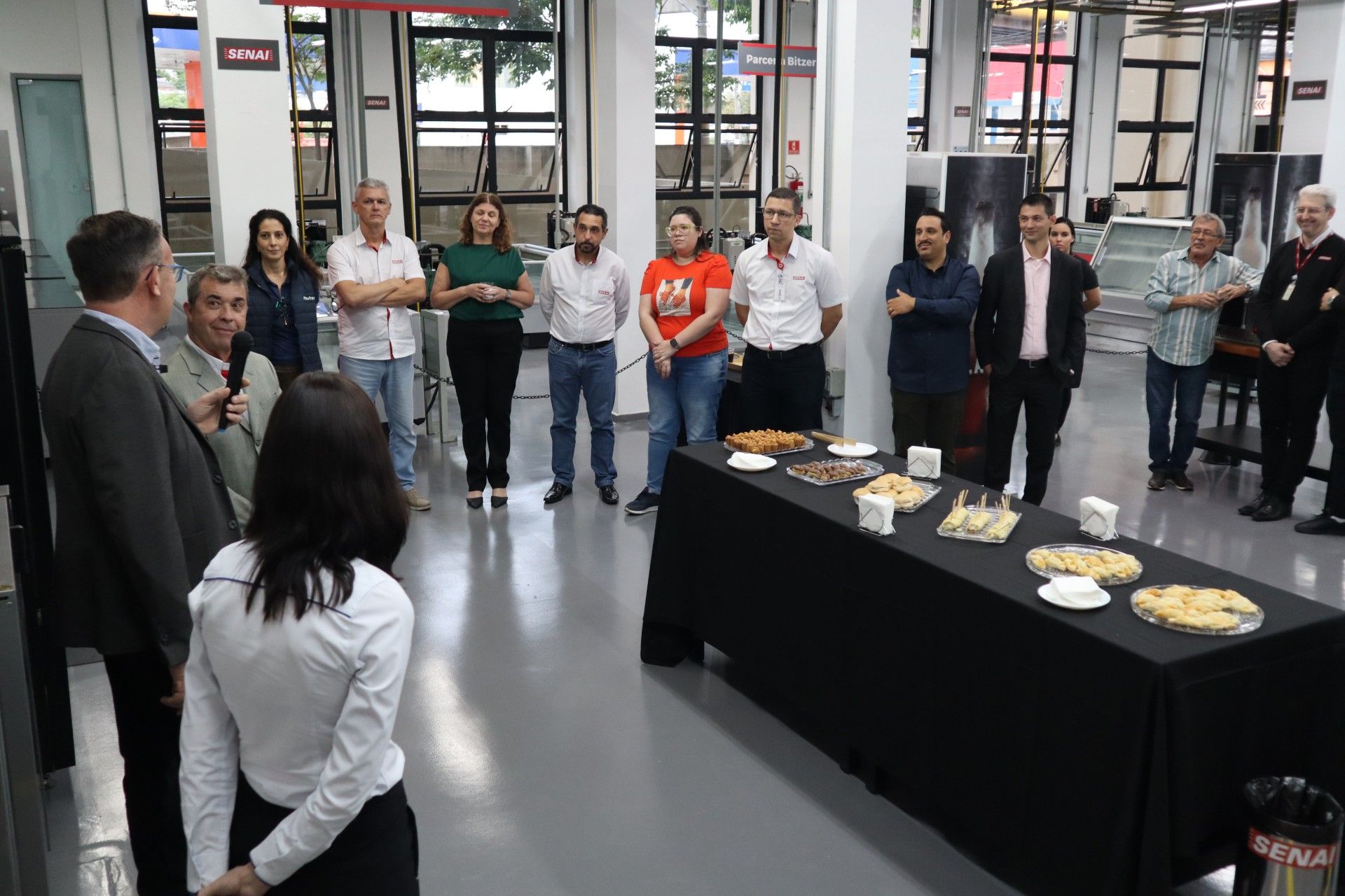 Pessoas reunidas em um ambiente interno, perto de uma mesa com comida, ouvindo um palestrante. Iluminação neutra; ambiente claro.
