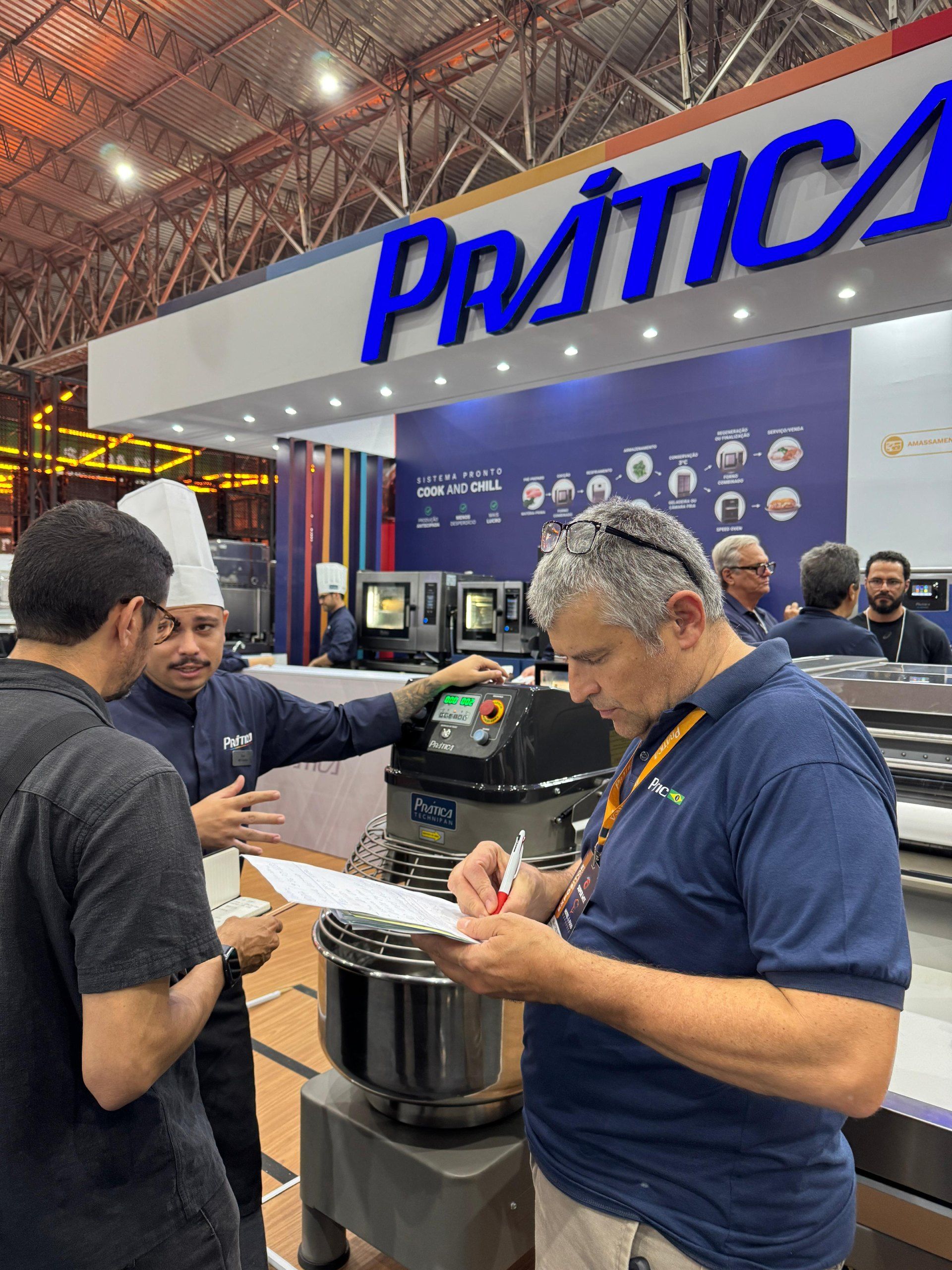Pessoas em um estande da Prática, uma delas anotando, um chef uniformizado explicando o funcionamento dos equipamentos em uma feira comercial.