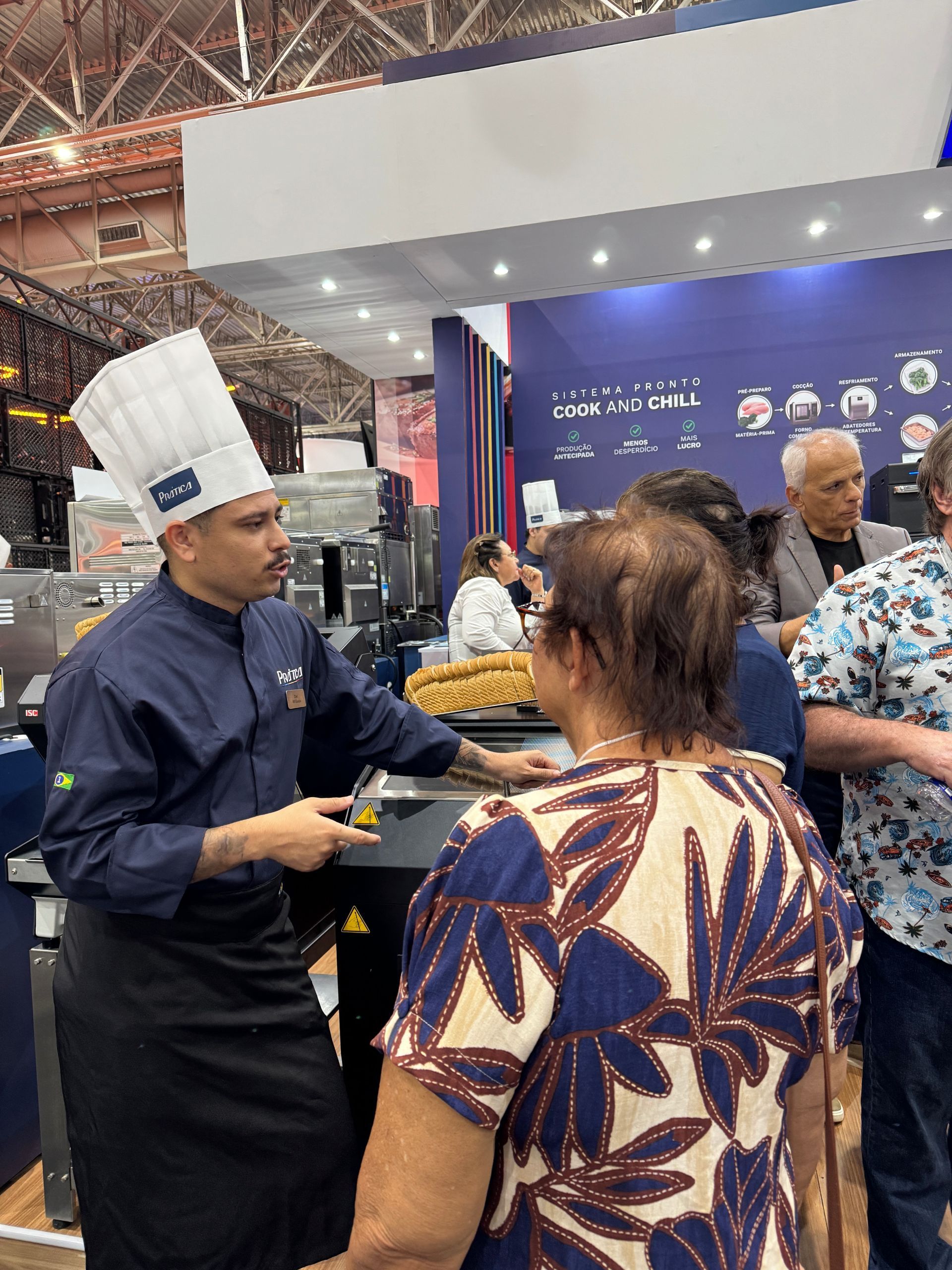Um chef uniformizado conversa com pessoas em uma barraca, segurando uma caneta. Ambiente de exposição, interior.