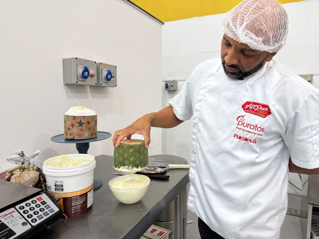 Um chef de jaleco branco examina um bolo decorado, a cozinha equipada com ingredientes e uma balança.