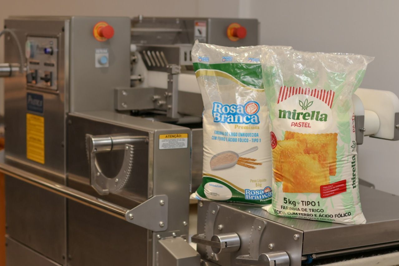 Sacos contendo produto alimentício branco ao lado de uma máquina de embalagem industrial. Rótulos visíveis.