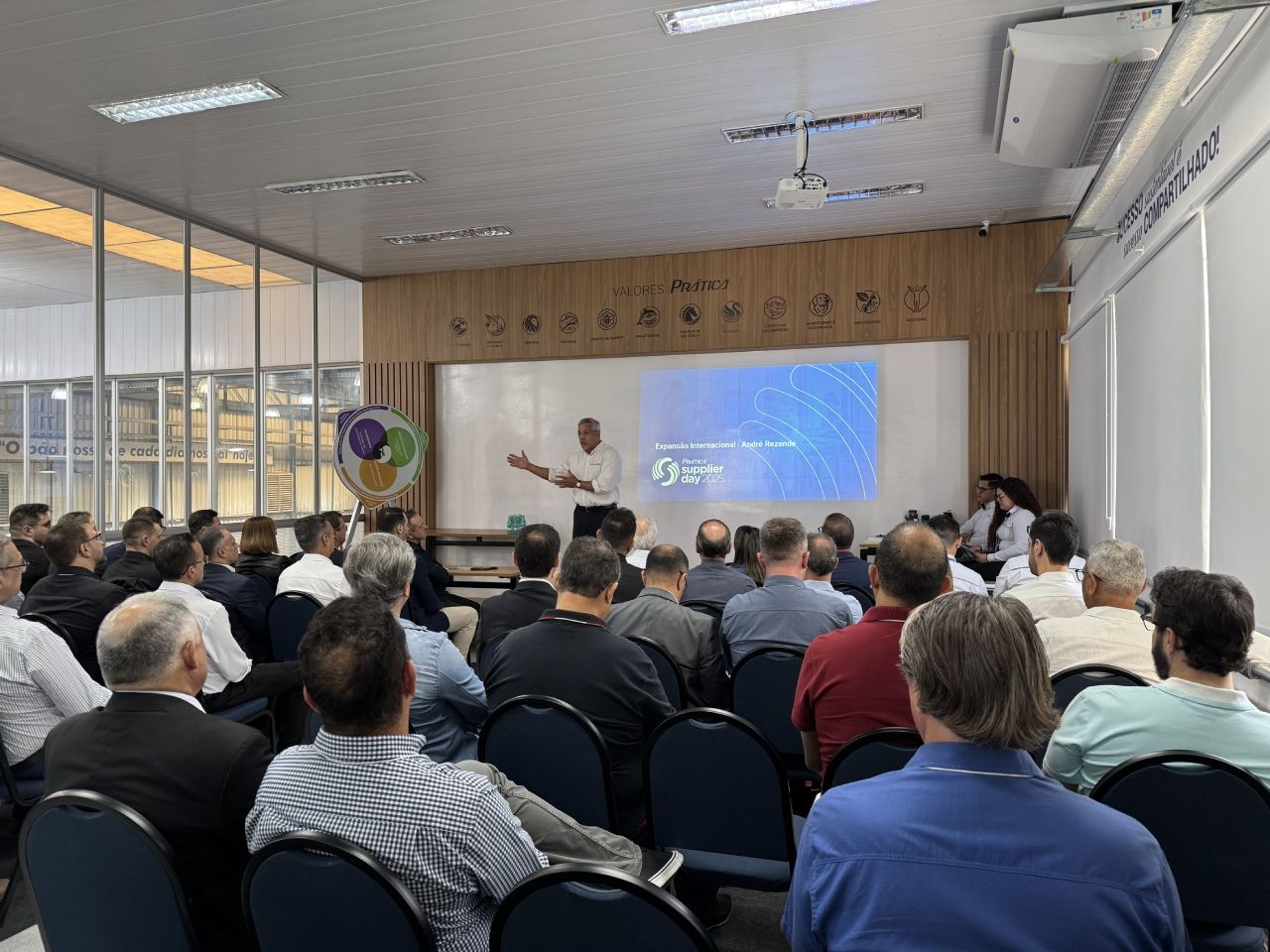 Homem fazendo apresentação em uma sala de aula para uma plateia sentada. Quadro branco com texto e imagens.