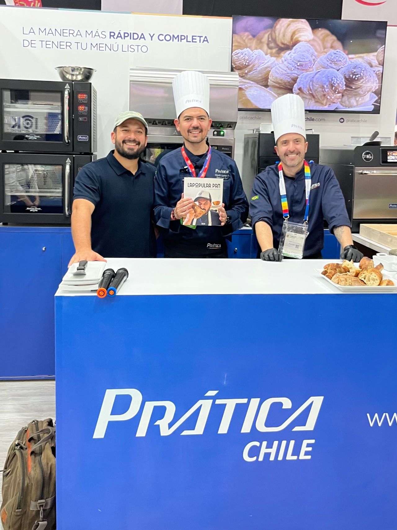 Três homens em um estande de exposição gastronômica, dois com chapéus de chef, um segurando um livro de receitas. Logotipo 