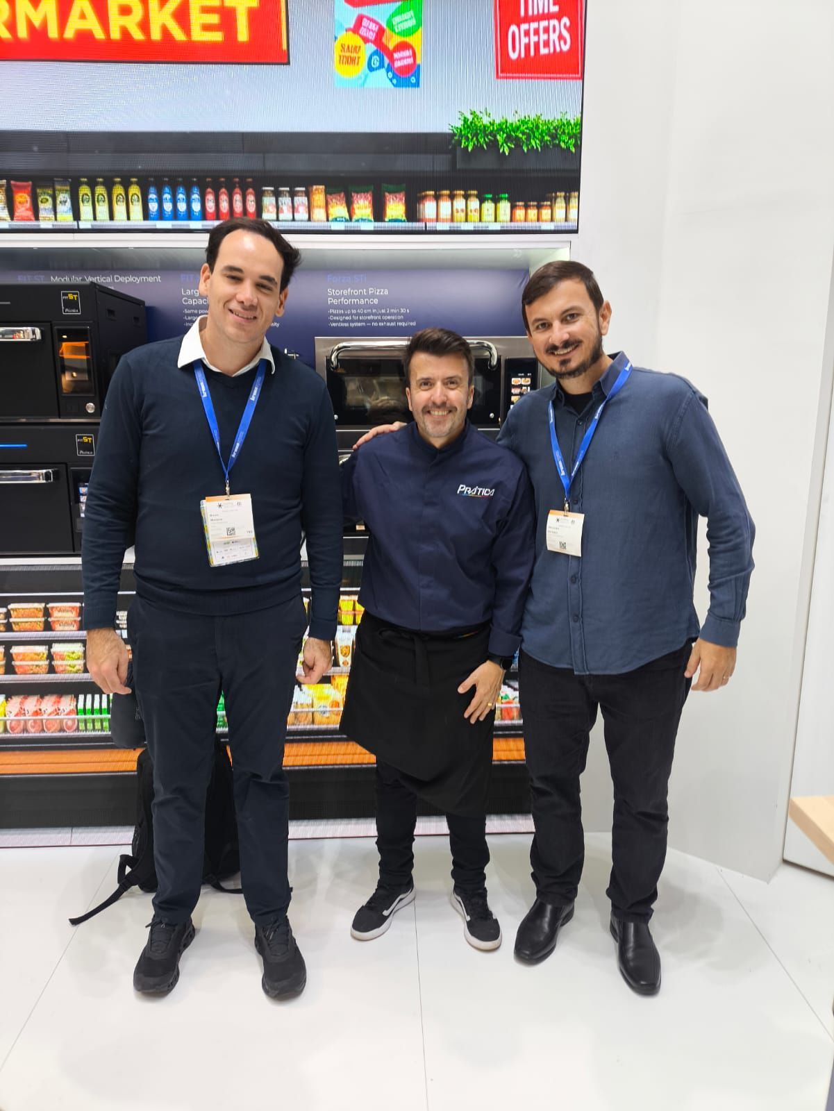 Três homens em uma feira comercial estão em frente a uma vitrine de supermercado. Um deles veste uniforme de chef.