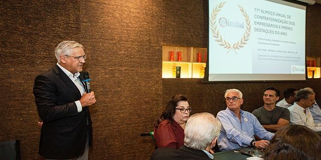 Um homem discursa em uma conferência. Várias pessoas ouvem atentamente, sentadas às mesas. Um telão exibe o texto. A sala tem uma parede escura.