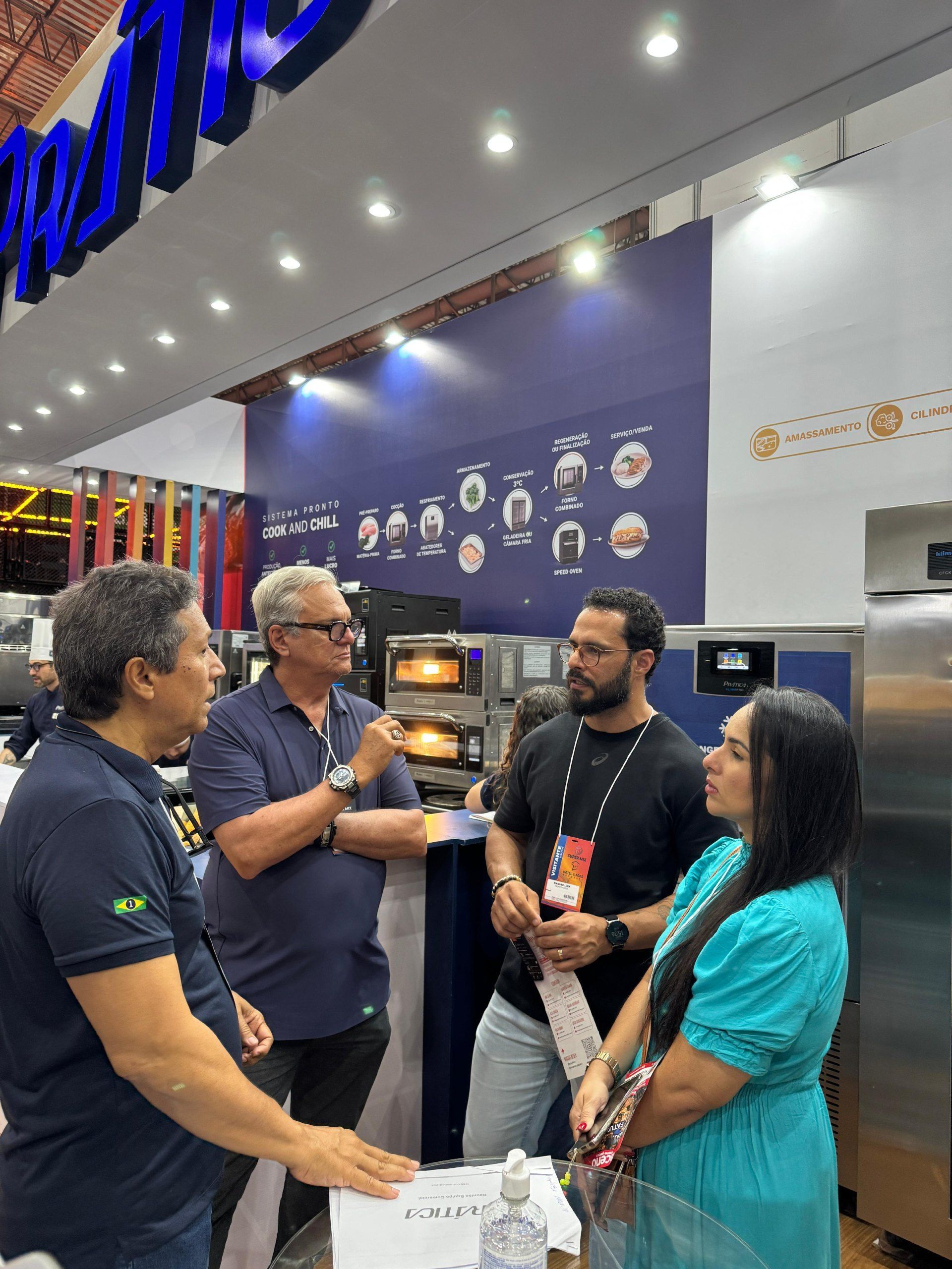 Quatro pessoas conversando em um estande de feira comercial perto de fornos de pizza. Parede azul com o logotipo da empresa visível.