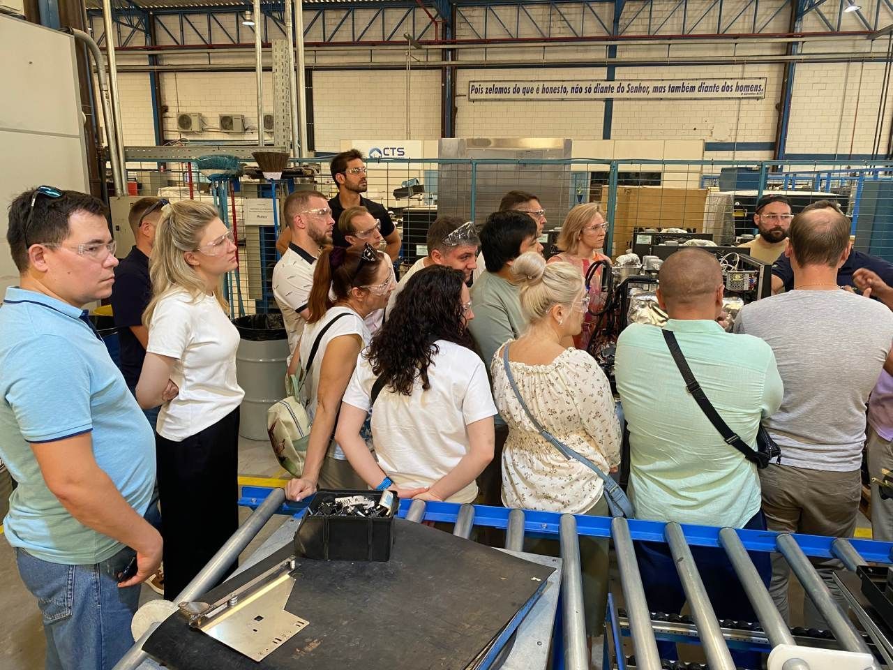 Um grupo de pessoas está em pé ao redor de uma correia transportadora em uma fábrica.