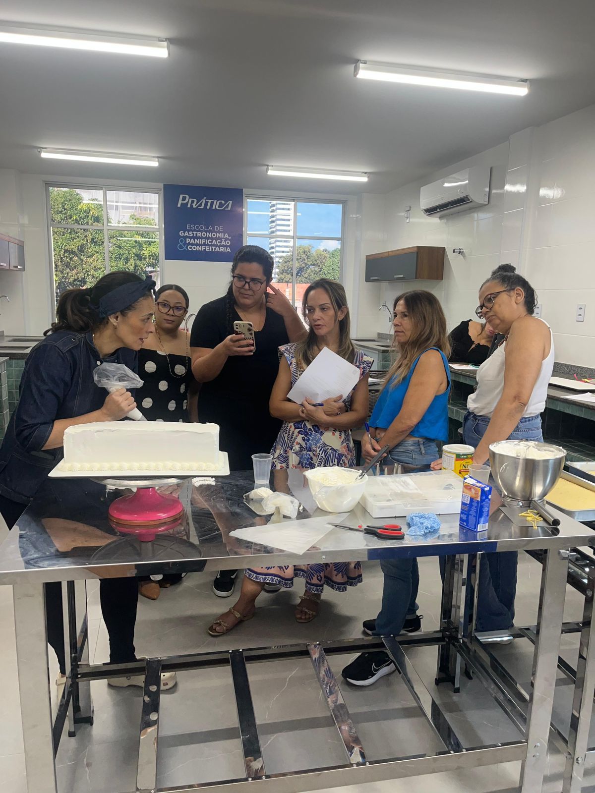 Uma aula de decoração de bolos: mulher alisando cobertura em um bolo, outras pessoas observando, em pé perto de uma mesa de aço inoxidável.