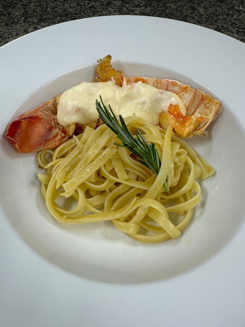 Cauda de lagosta com molho cremoso e massa fettuccine, decorada com alecrim em prato branco.